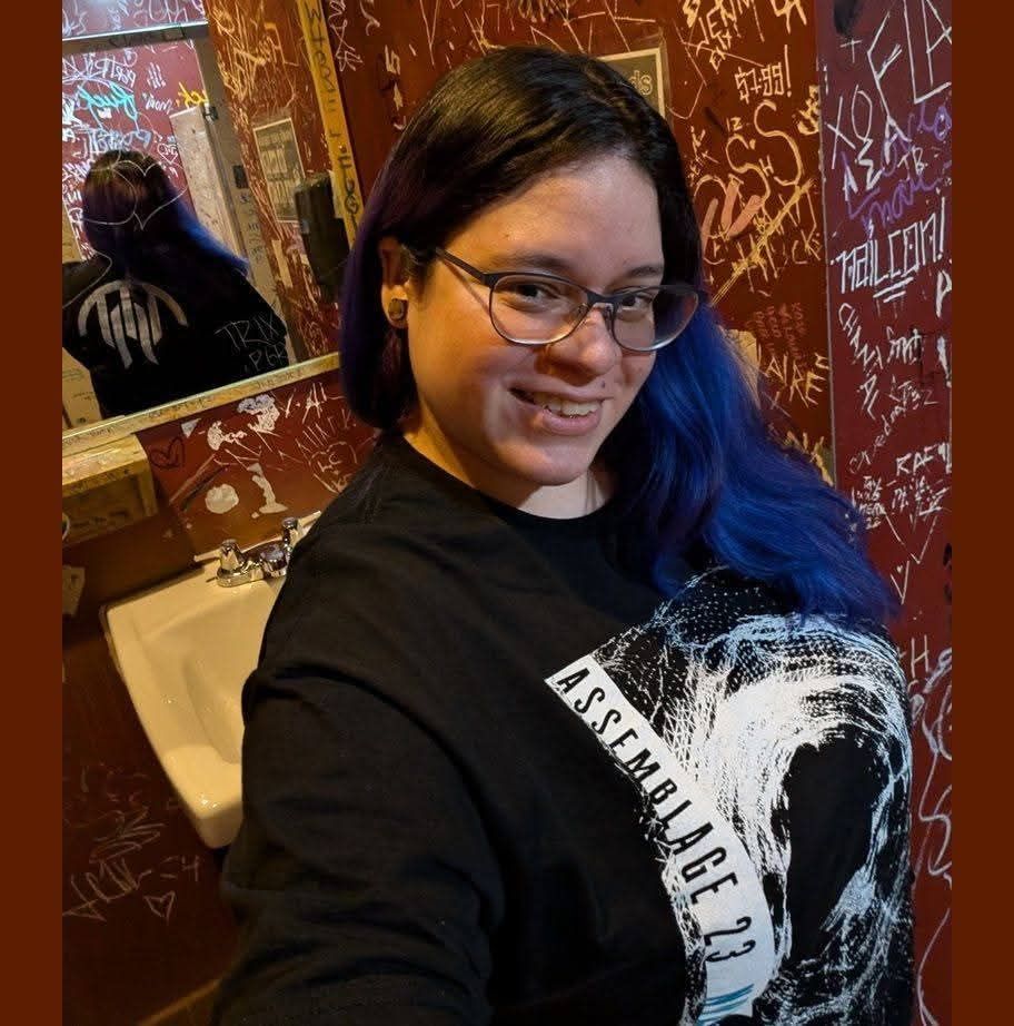 Person taking a mirror selfie in a restroom, wearing glasses and a black shirt with blue hair.