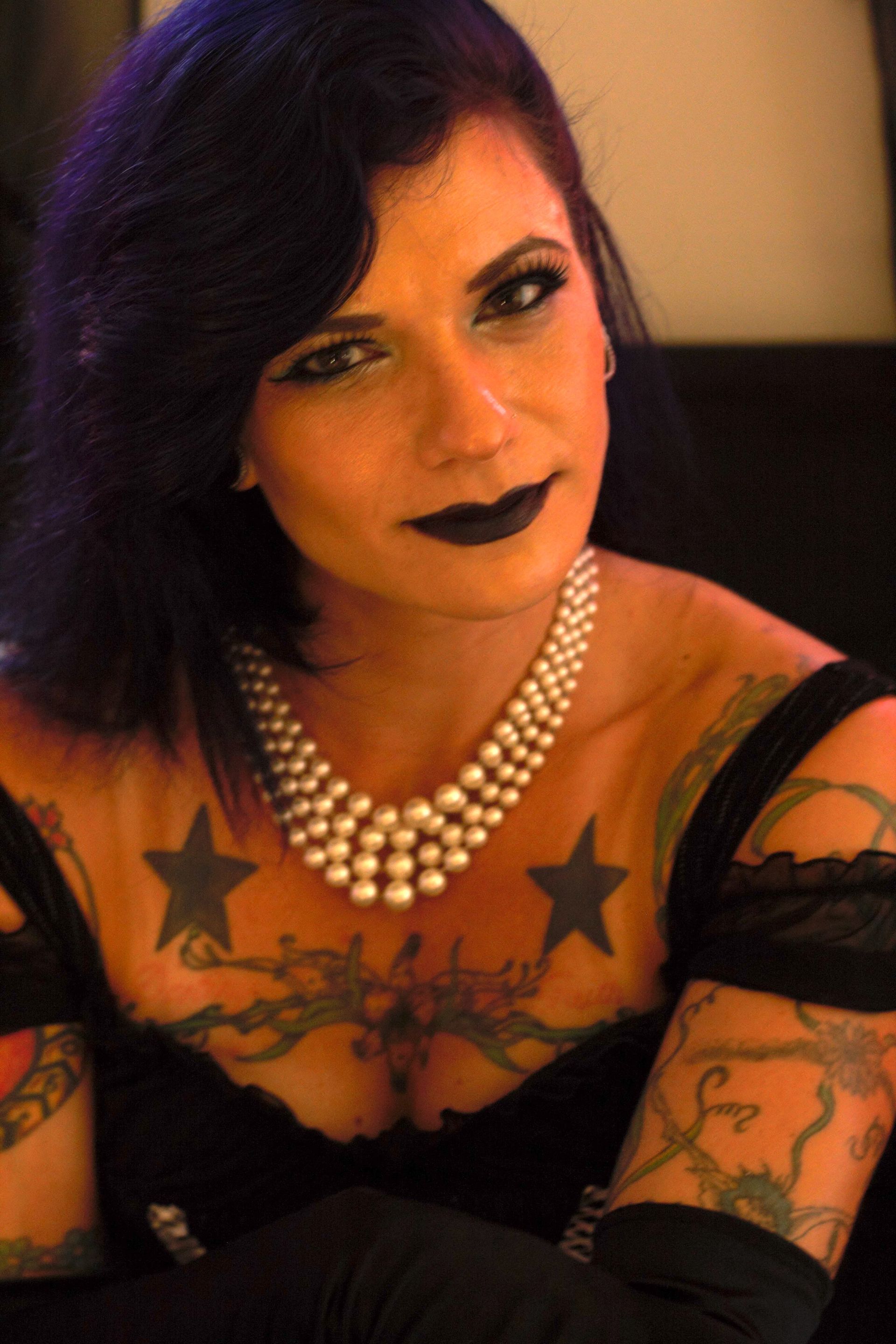 Woman with dark hair and hoop earrings, posing indoors in a black top with tattooed arms, abstract art behind her