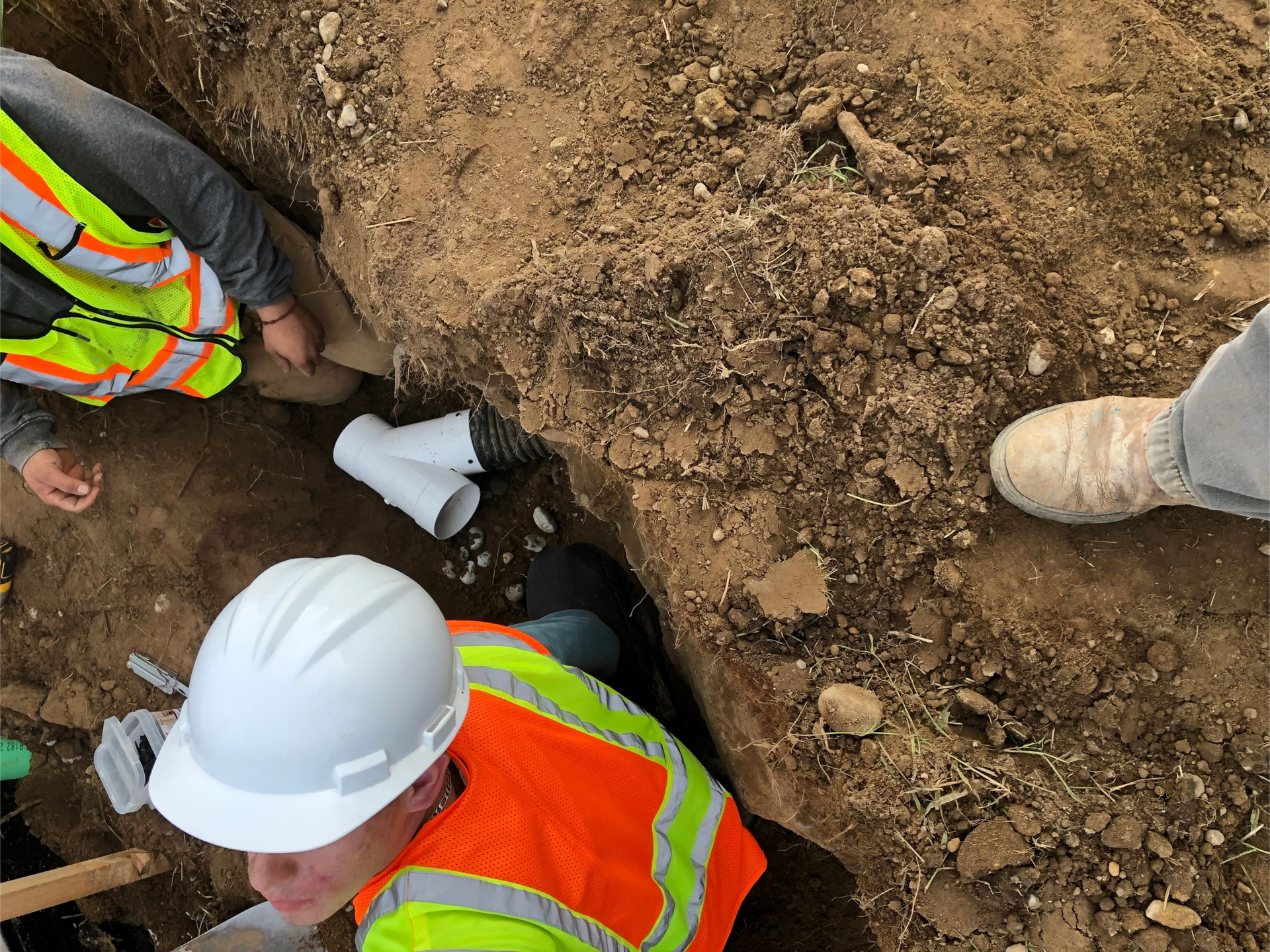 Construction workers in a trench installing pipes. One wears a hard hat and safety vest.