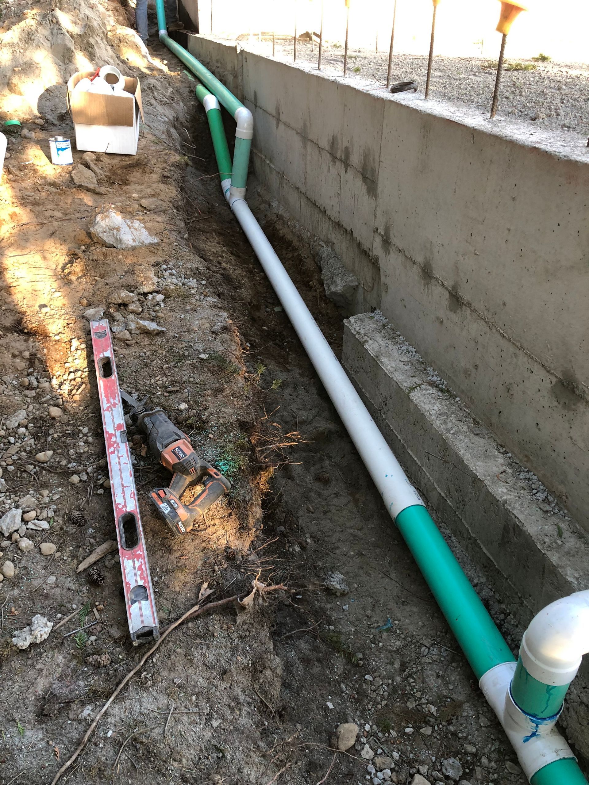 Installation of green and white plumbing pipes next to a concrete wall in a trench.