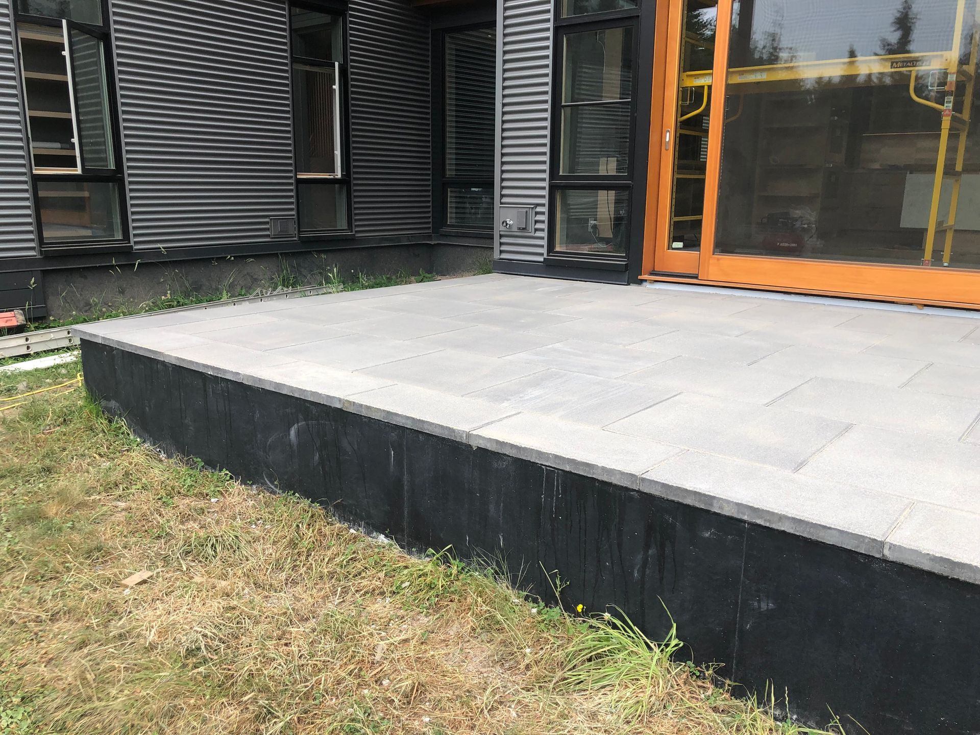 Gray paver patio in front of a modern building, black base, grass foreground.