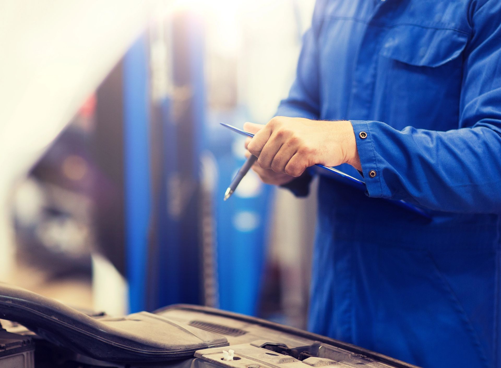 Meccanico in tuta blu, con blocco per appunti in mano, ispeziona il motore di un'auto. Ambiente da officina.