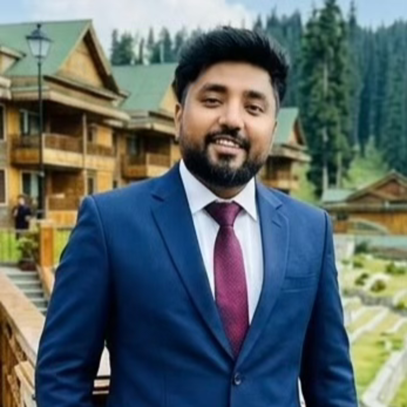 Man in a blue suit and maroon tie smiling outdoors with wooden cabins and pine trees in the background