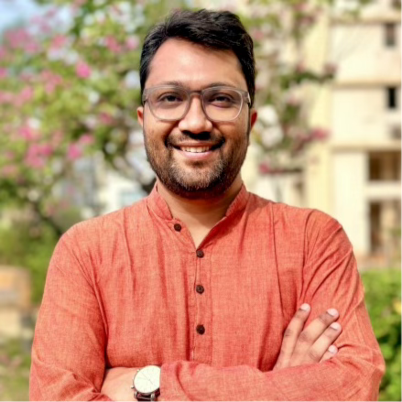 Smiling person in an orange shirt with arms crossed, standing outdoors with blurred trees and buildings behind.