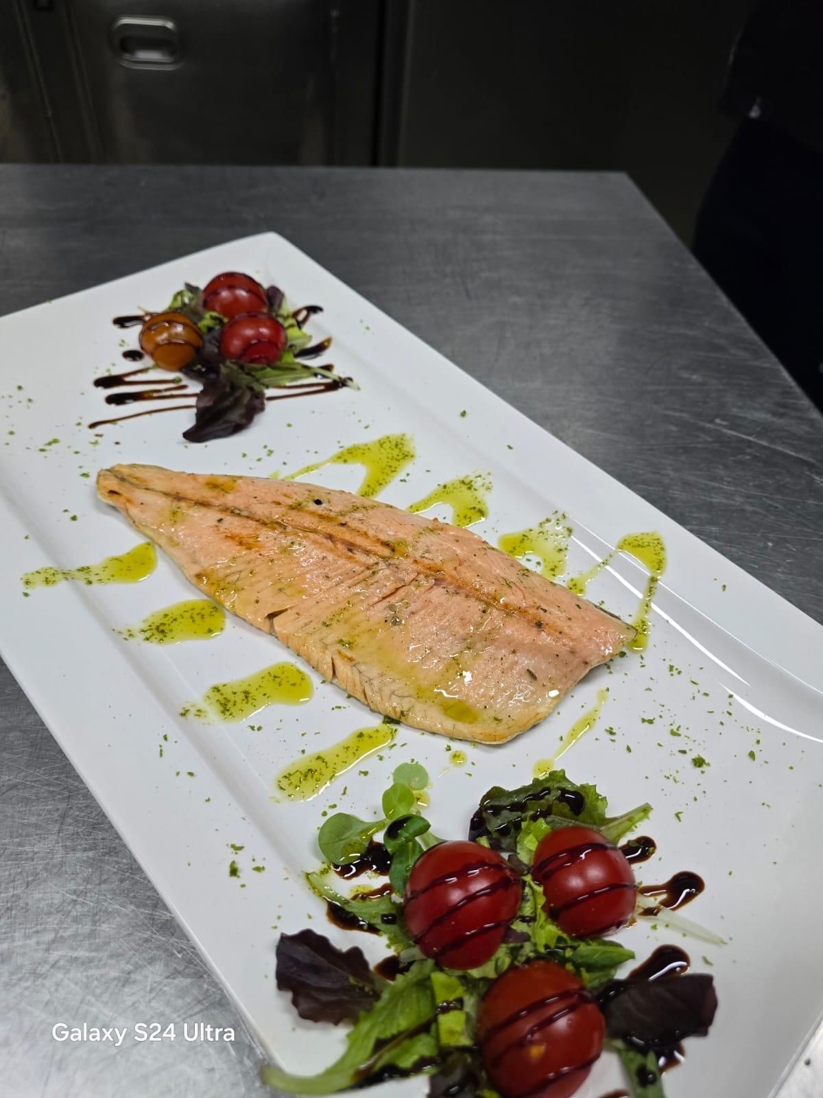 Filetto di salmone grigliato su un piatto bianco, guarnito con pomodorini, verdure e un filo d'olio.