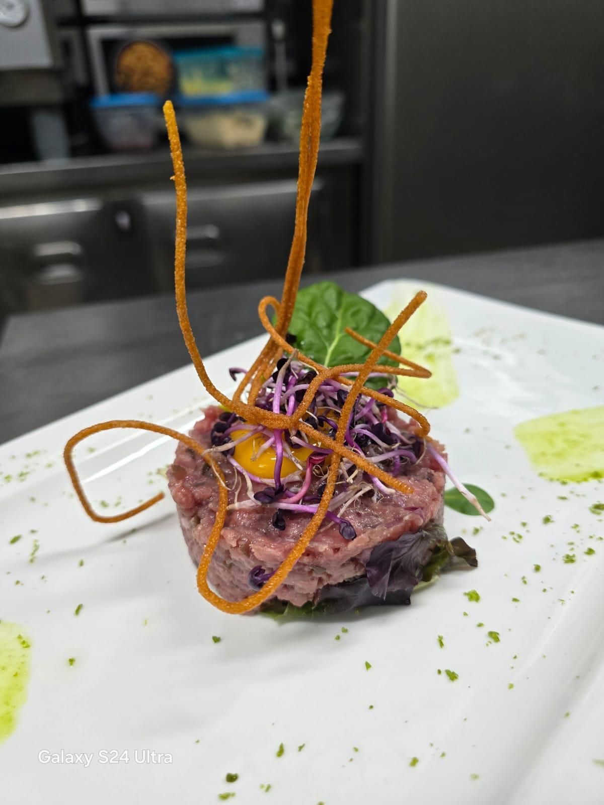 Antipasto: Tartare di carne, guarnita con germogli, filetti fritti e salsa verde su un piatto bianco.