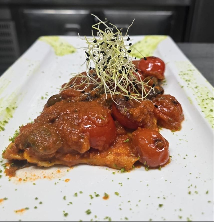 Piatto di pesce impanato con salsa di pomodoro e pomodorini, guarnito con germogli, su un piatto rettangolare.