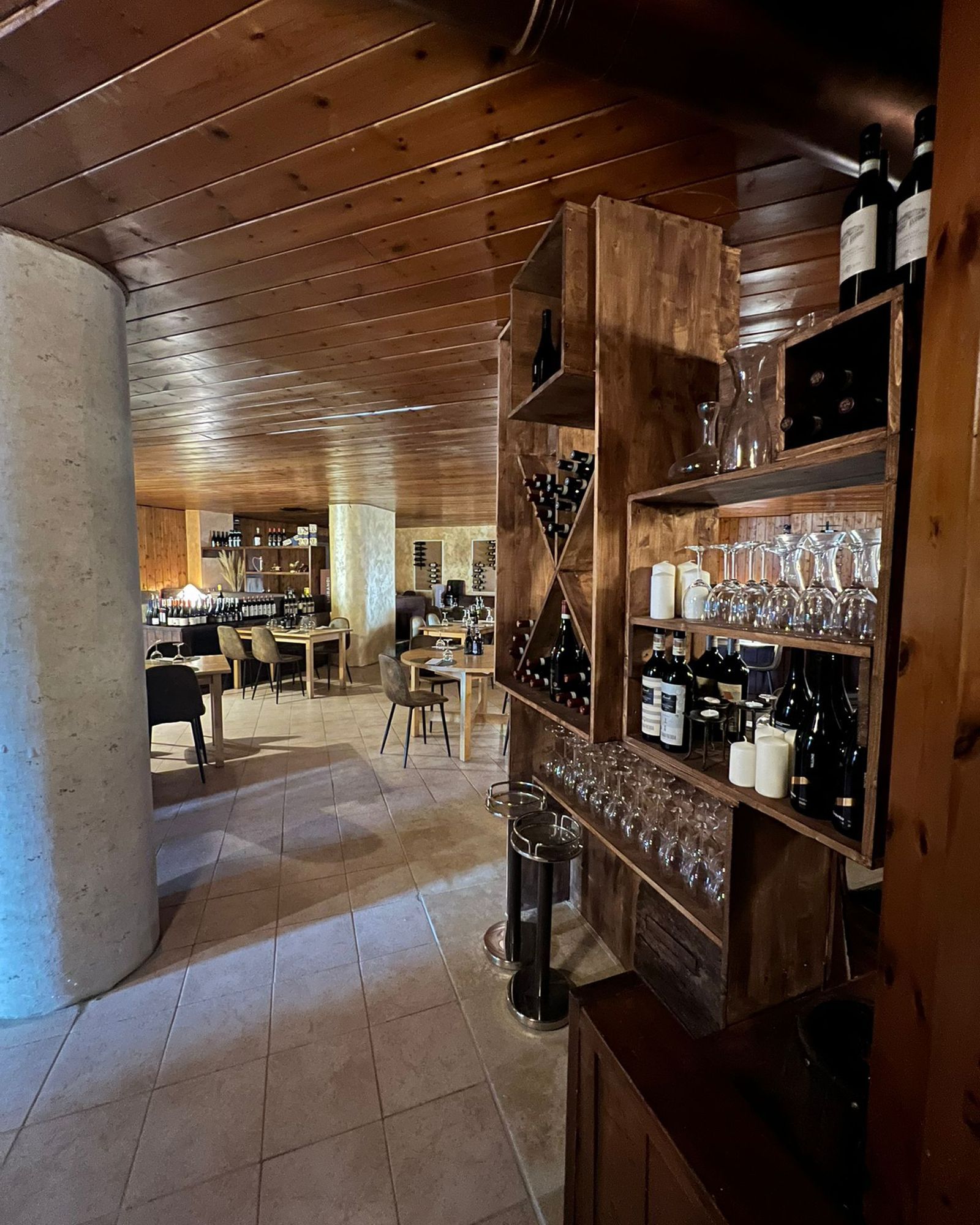 Interno del ristorante con portabottiglie, bicchieri e tavoli con sedie. Soffitto e pavimento in legno.