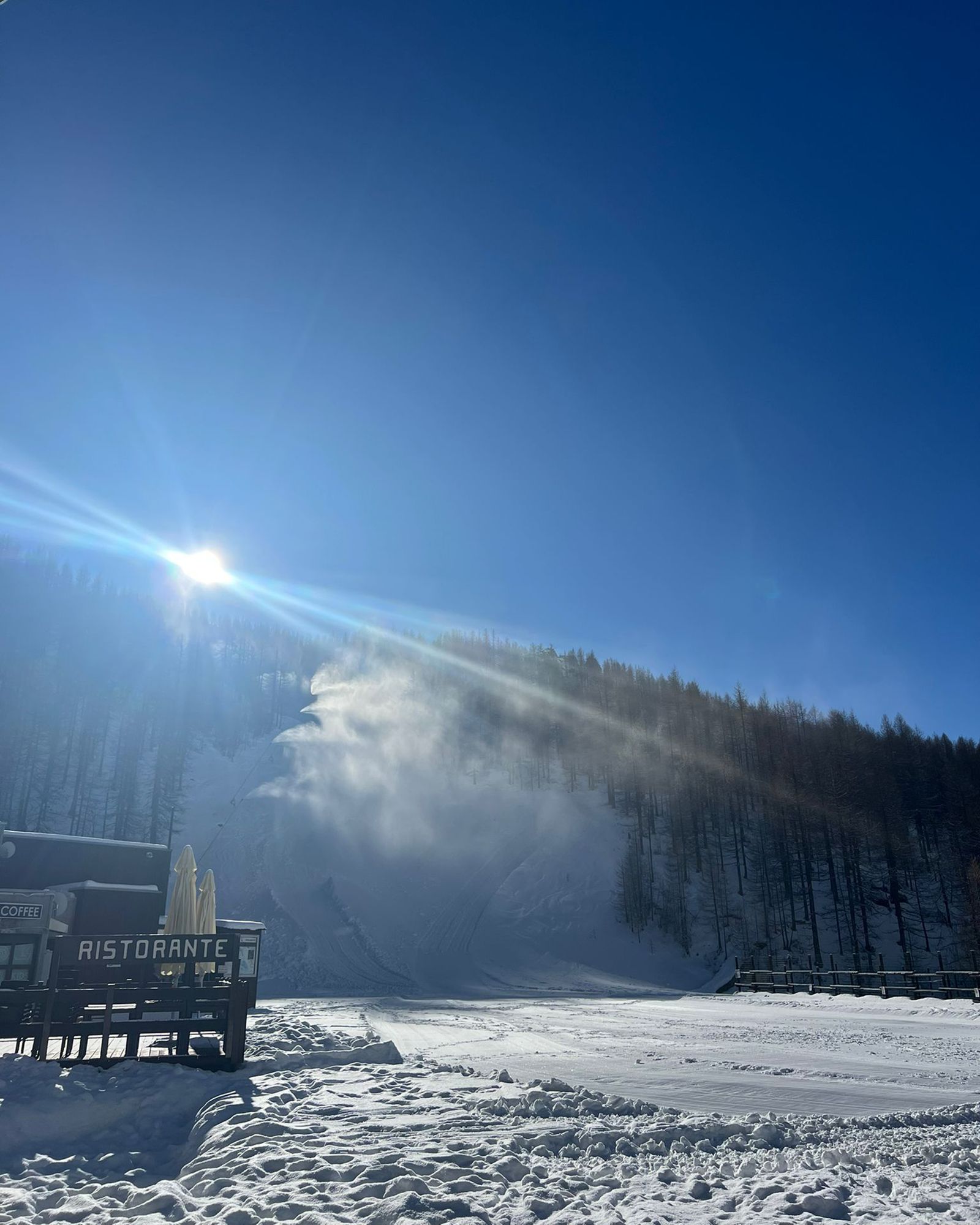 Luce del sole su un paesaggio innevato con un edificio sulla sinistra, una foresta sulla destra e un cielo azzurro brillante.