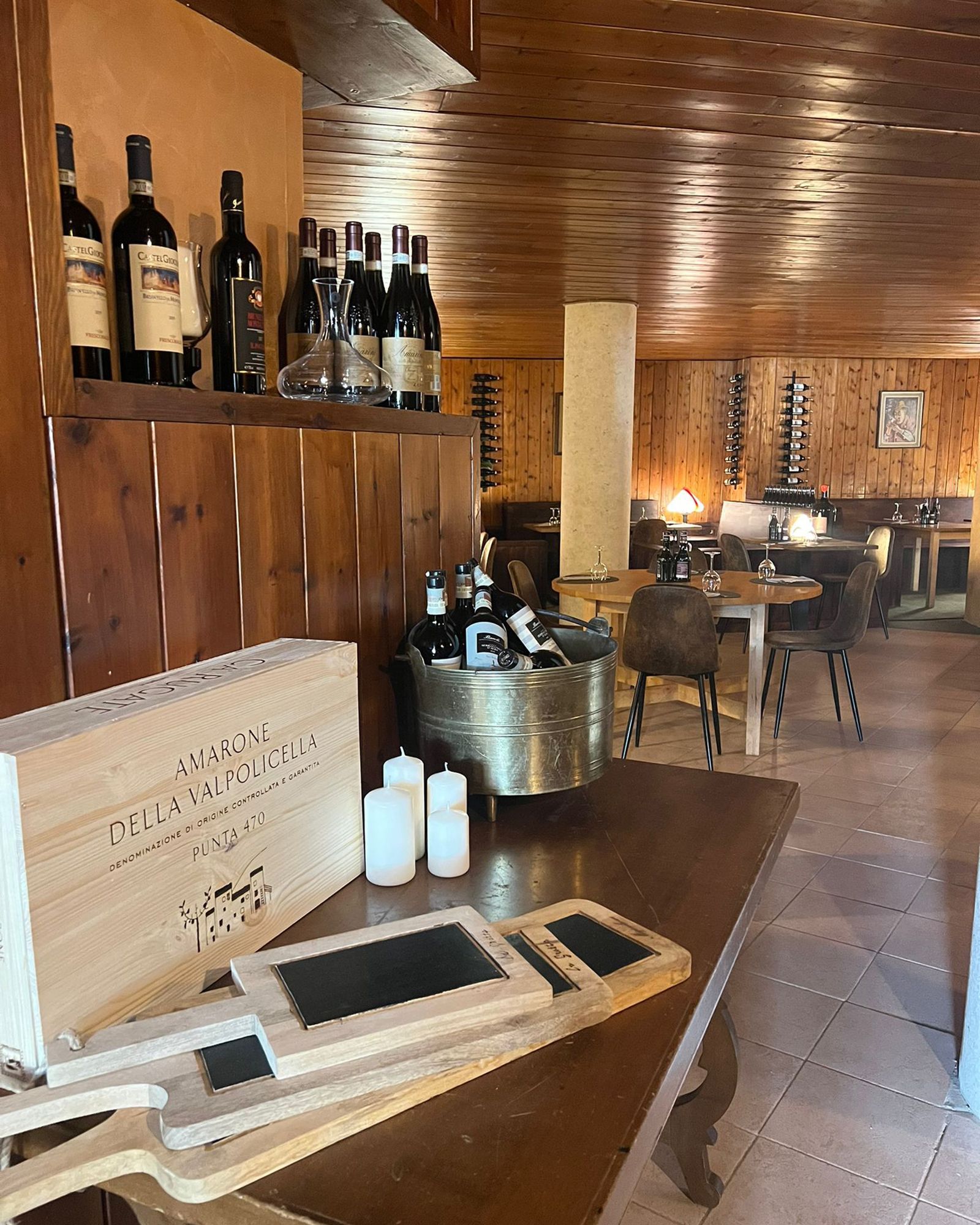 Bottiglie di vino sugli scaffali e in un secchio di metallo all'interno di un ristorante con pannelli in legno. Di fronte, un tavolo di legno con una cassa di legno e delle lavagne.