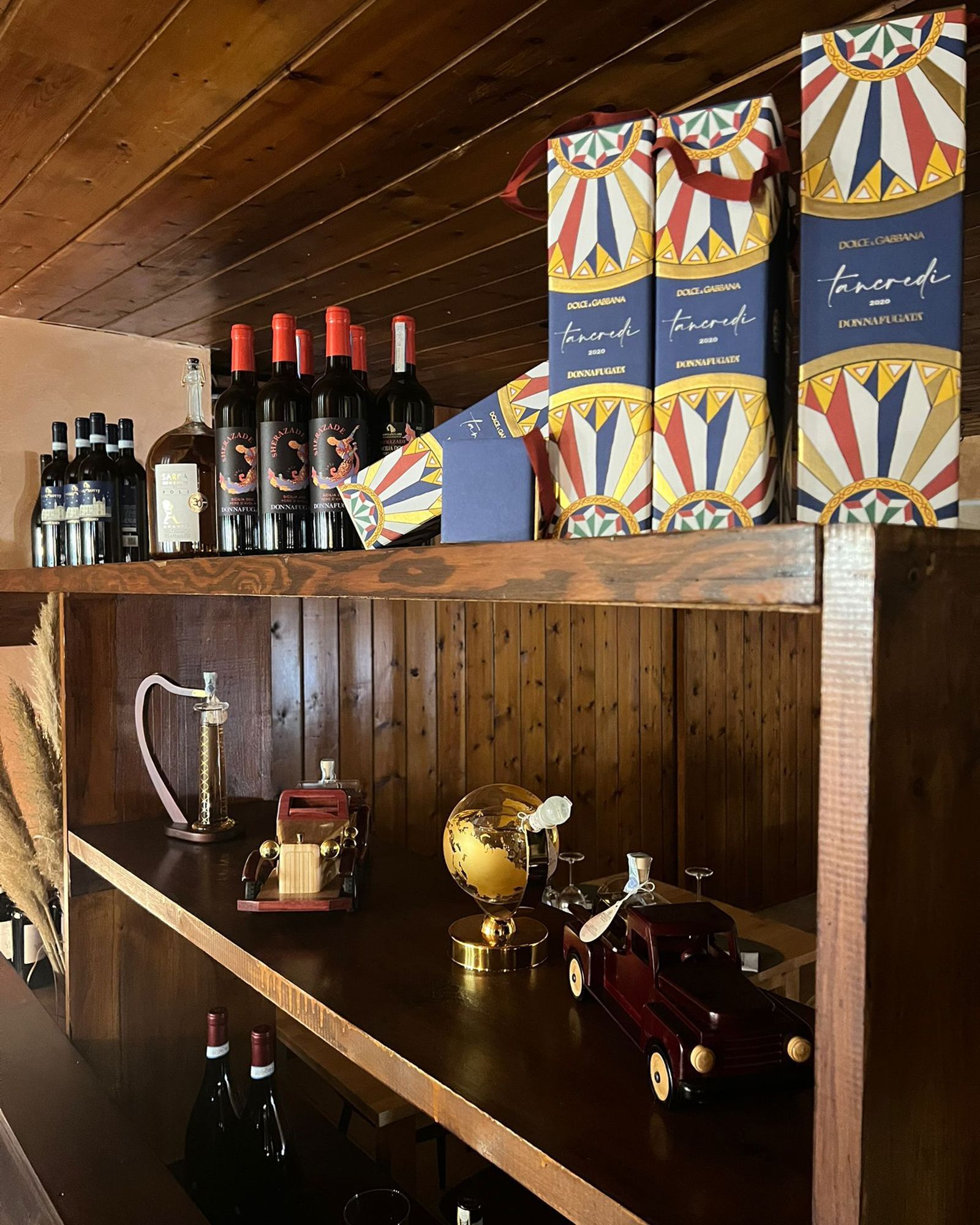 Scaffali in legno su cui sono esposte bottiglie di vino, scatole regalo e oggetti decorativi, in un interno rustico e scarsamente illuminato.