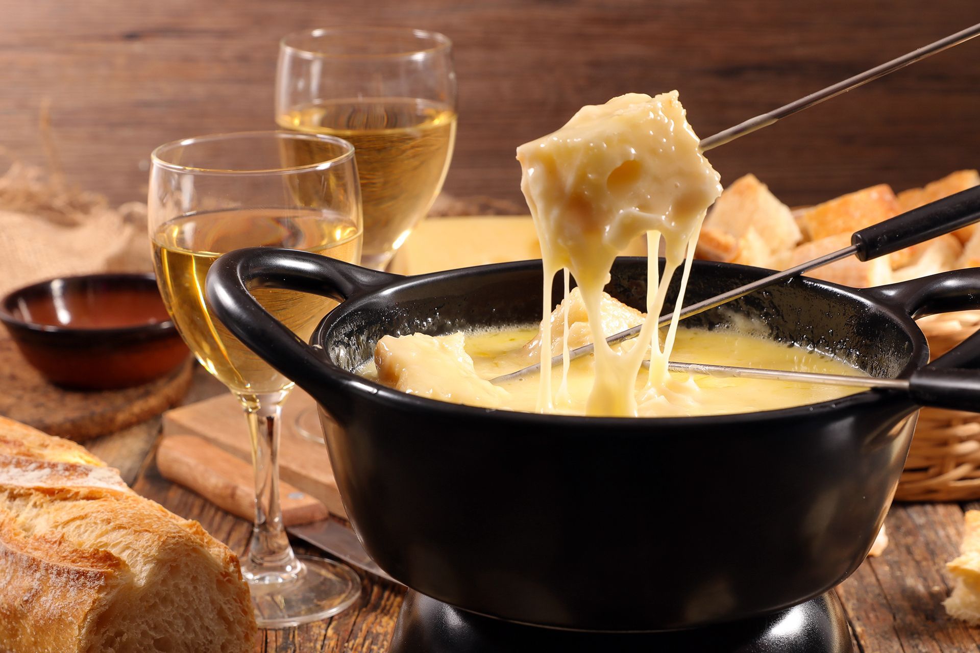 Fonduta di formaggio in una pentola nera, pane sulle forchette, bicchieri da vino bianco e fette di pane su un tavolo di legno.