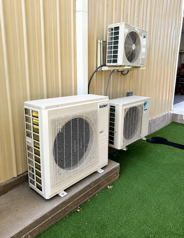Three outdoor air conditioner units beside a beige wall, with grass turf below.