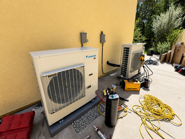 Outdoor HVAC equipment beside a yellow stucco wall, with hoses and tools on the ground.