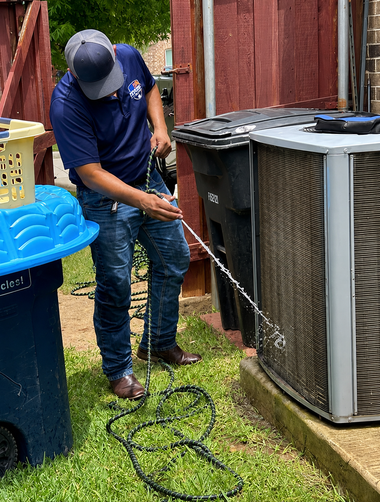 Worker pressure-washing a large outdoor HVAC unit beside a fence and bin in a backyard
