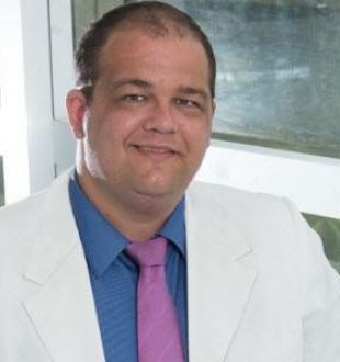 Homem sorrindo, usando terno branco, camisa azul e gravata rosa, em ambiente interno iluminado.