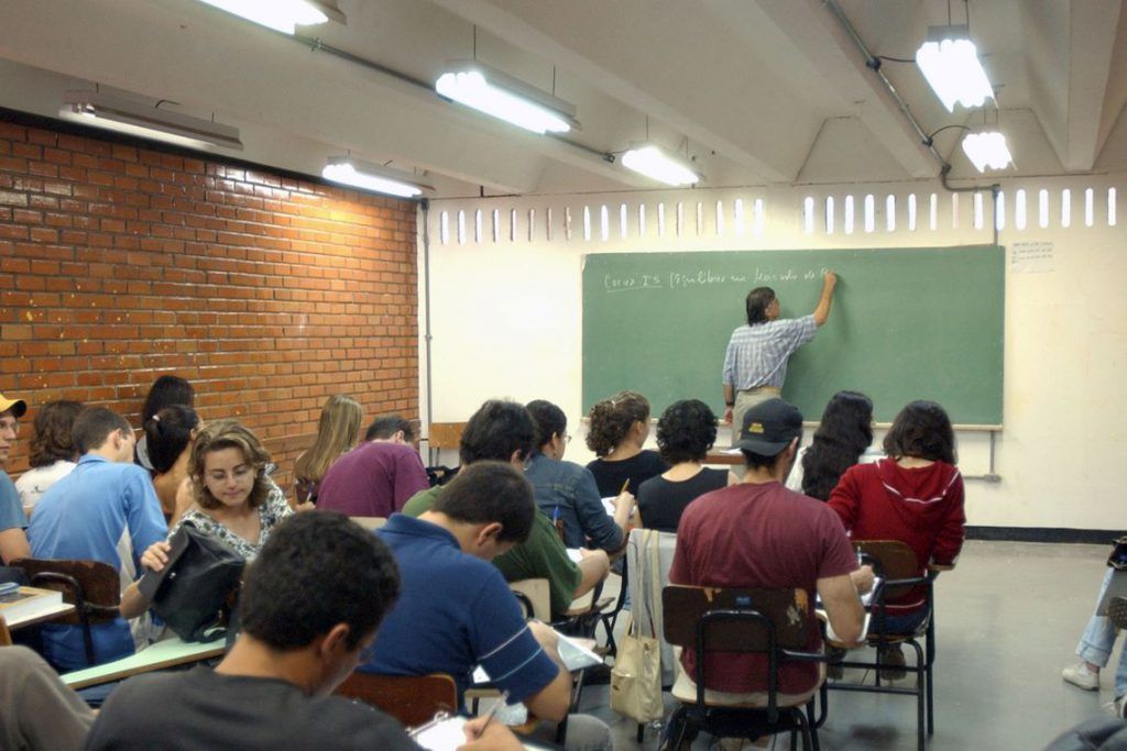 Alunos em sala de aula prestando atenção em aula com professor no quadro.