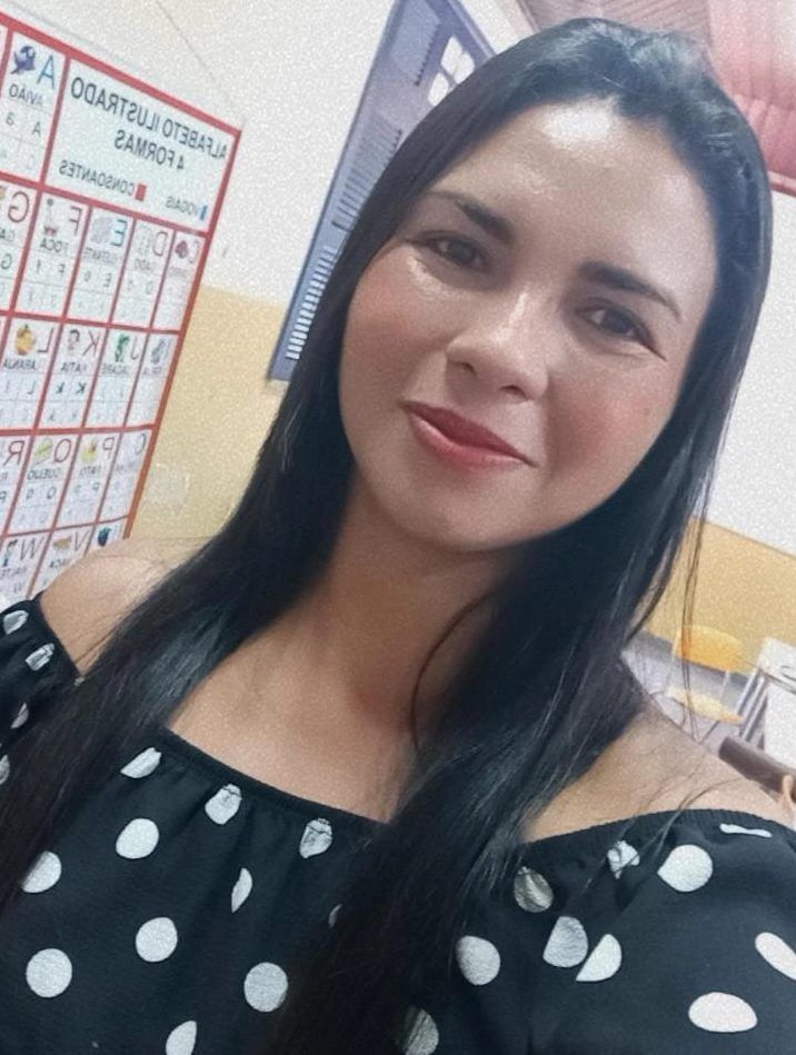 Mulher sorri em selfie tirada em sala de aula, usando blusa preta com bolinhas brancas.