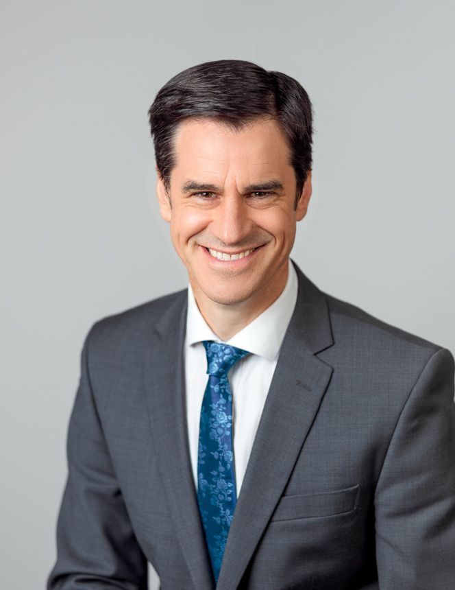 Retrato de Luke Davies, usando terno cinza e gravata azul, sorrindo diante de fundo cinza claro.
