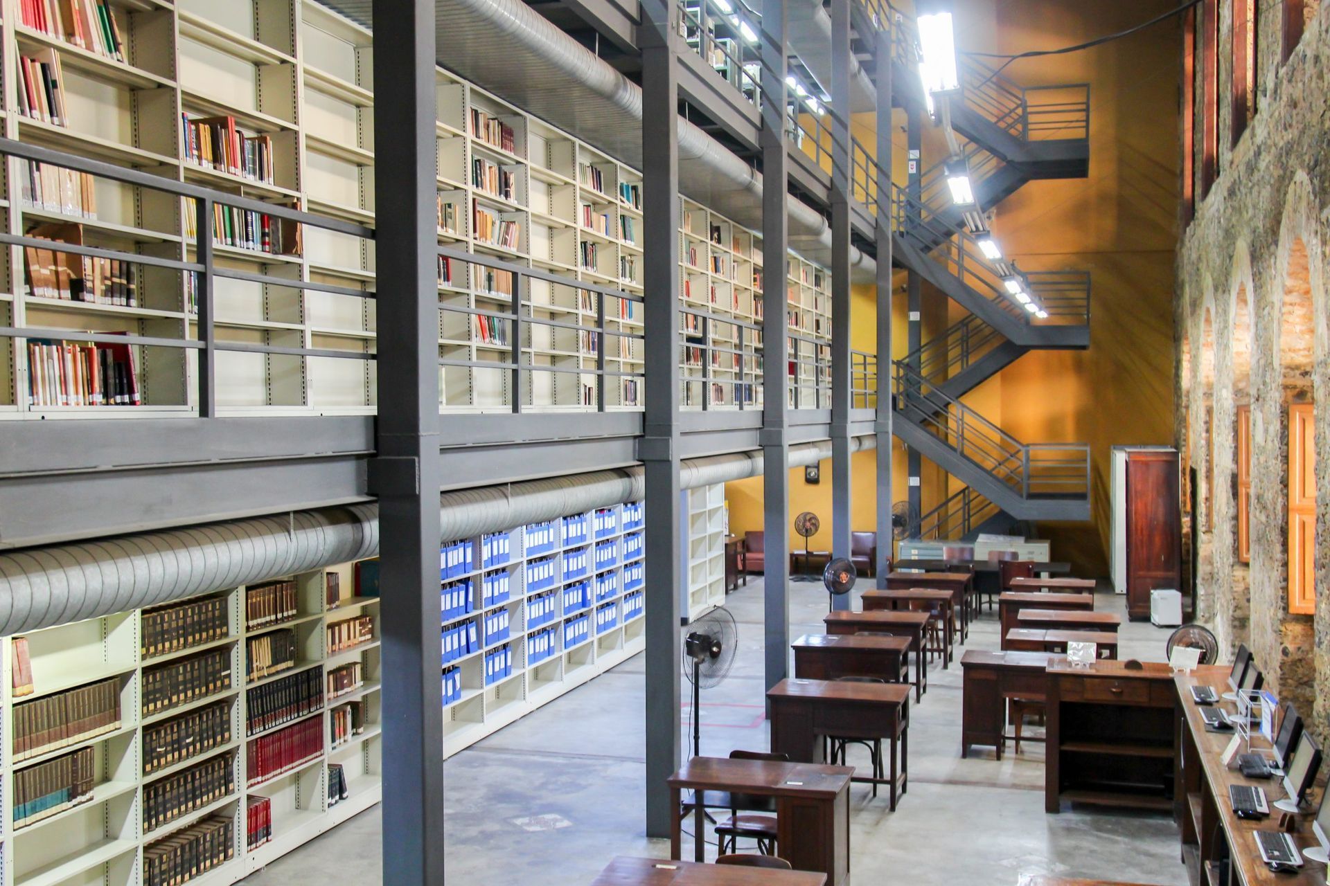 Biblioteca ampla com estantes de livros, escadas metálicas e mesas de estudo individuais.