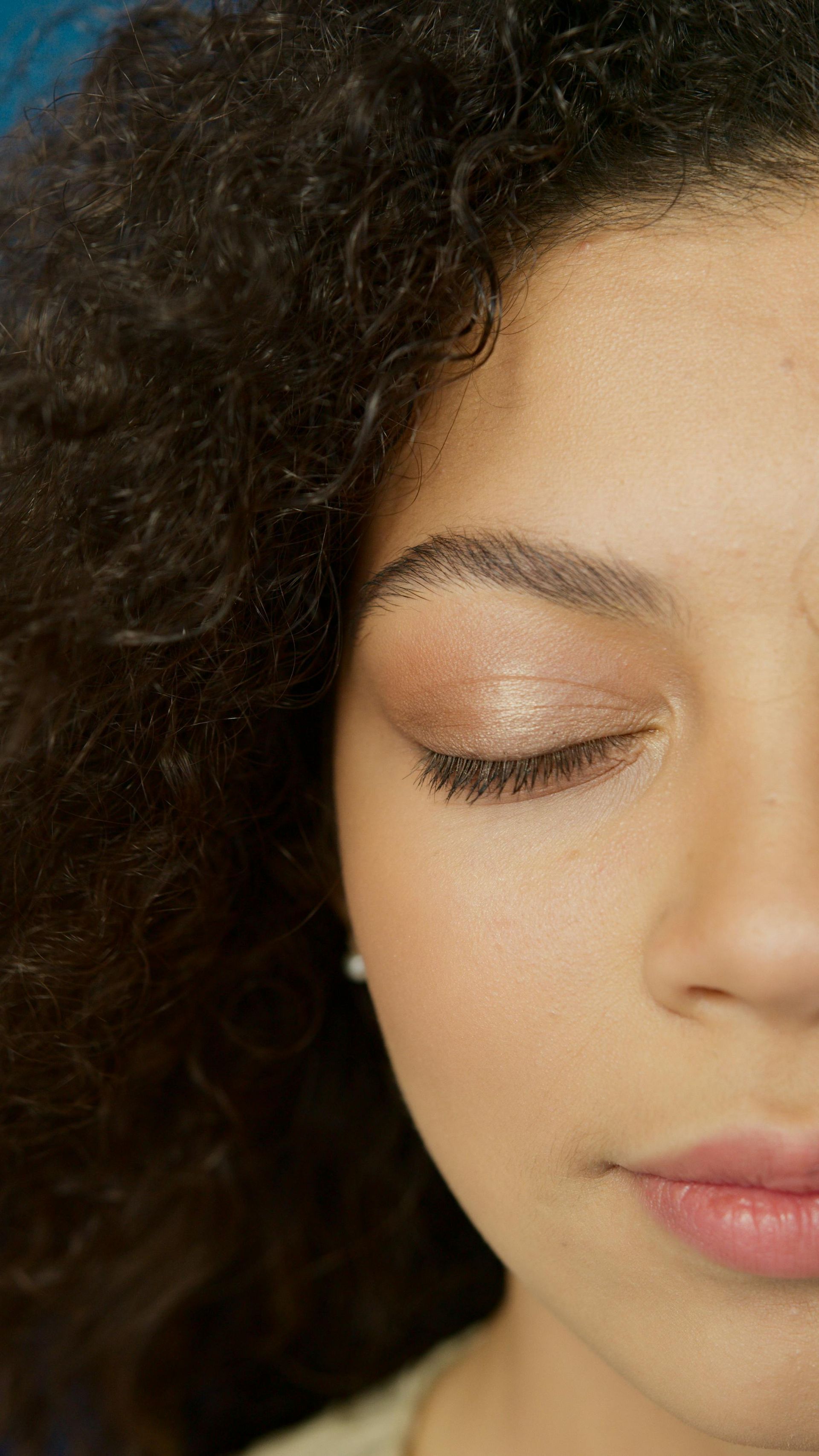 Close-up of a Person With Curly Dark Hair, Eyes Closed, Wearing Soft Neutral Makeup — Miilan Hair in Darwin City, NT