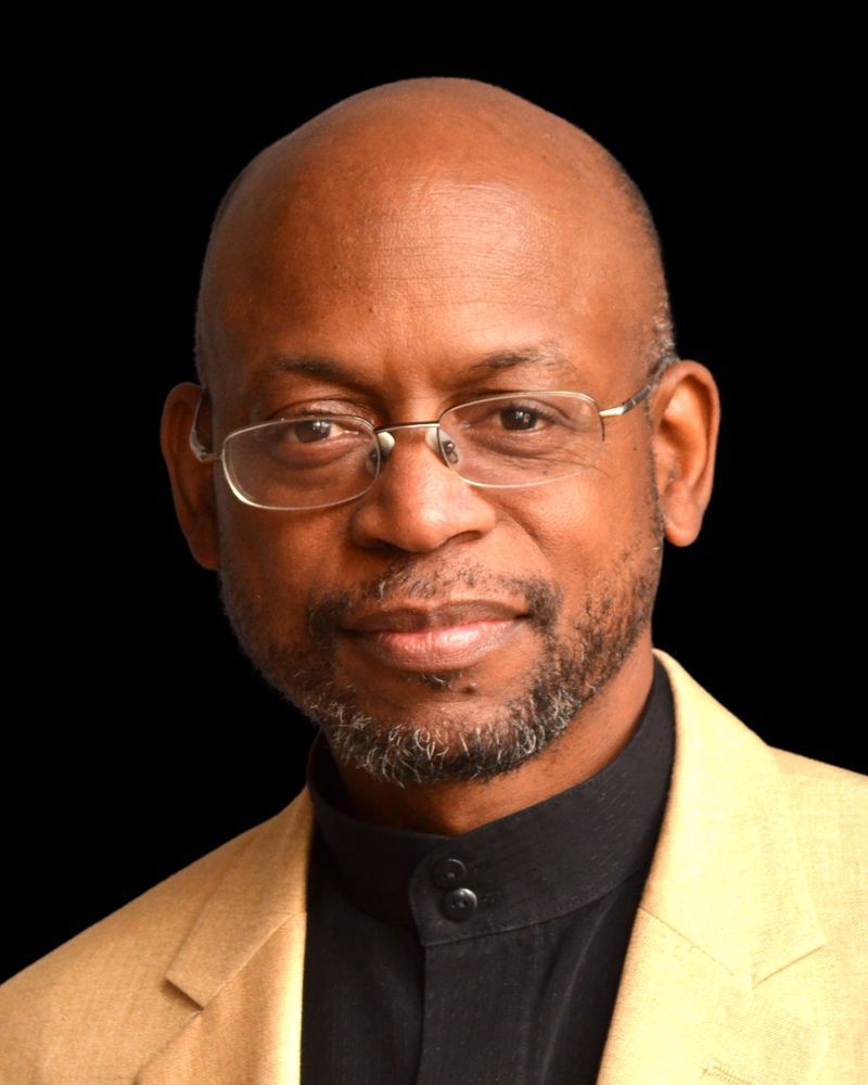 Bald man with glasses wearing a tan jacket and black shirt, smiling at the camera.