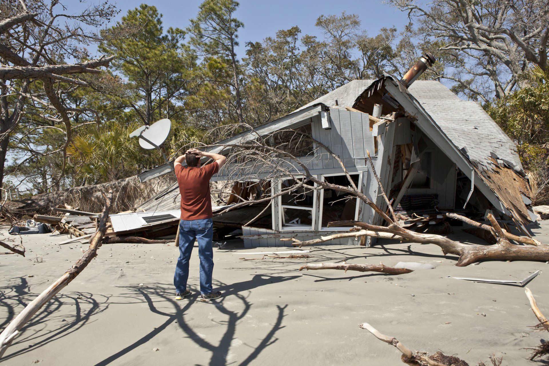 Man stands before a partially destroyed house on a sandy beach.