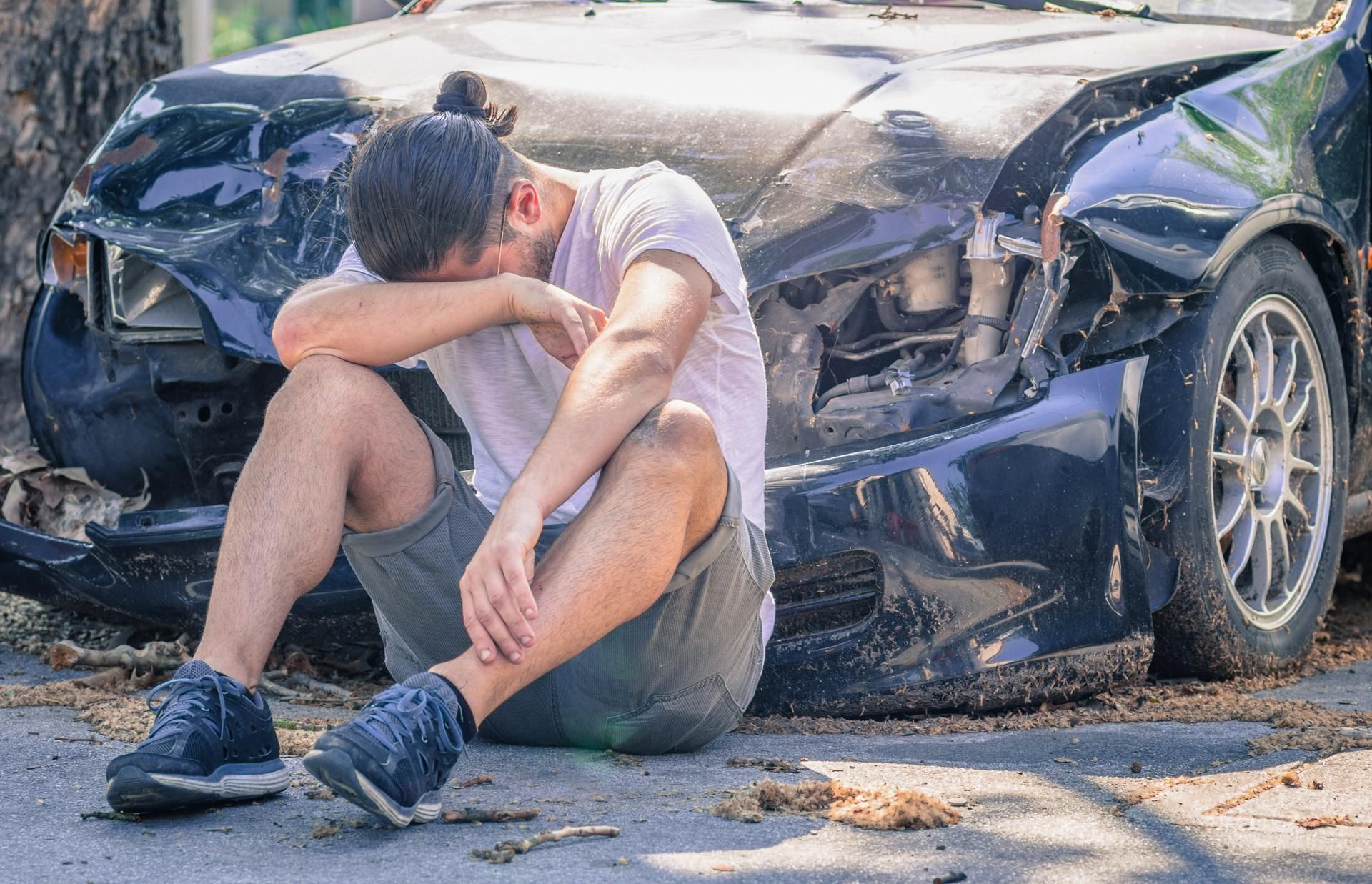A man is sitting on the ground in front of a damaged car.