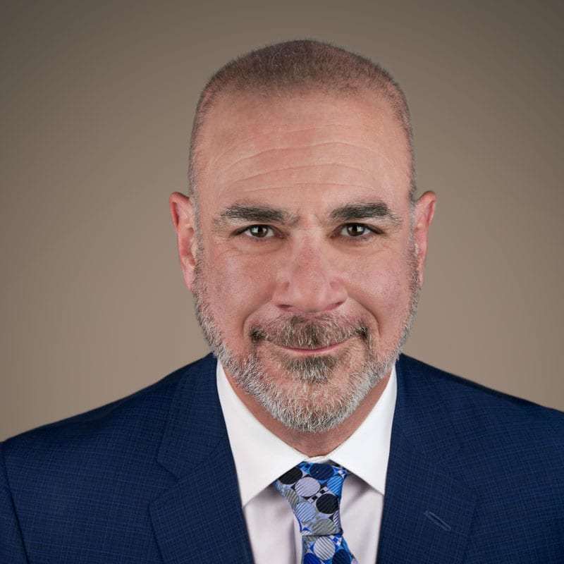 Man in blue suit, smiling slightly, against a neutral background. White shirt, patterned tie.
