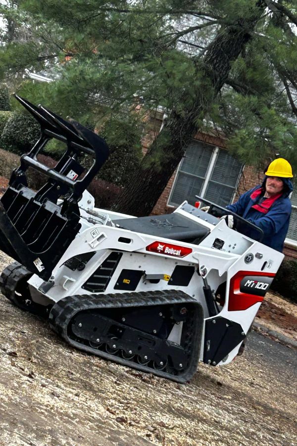 A person wearing a yellow hard hat operates a white and red Bobcat compact track loader outdoors.