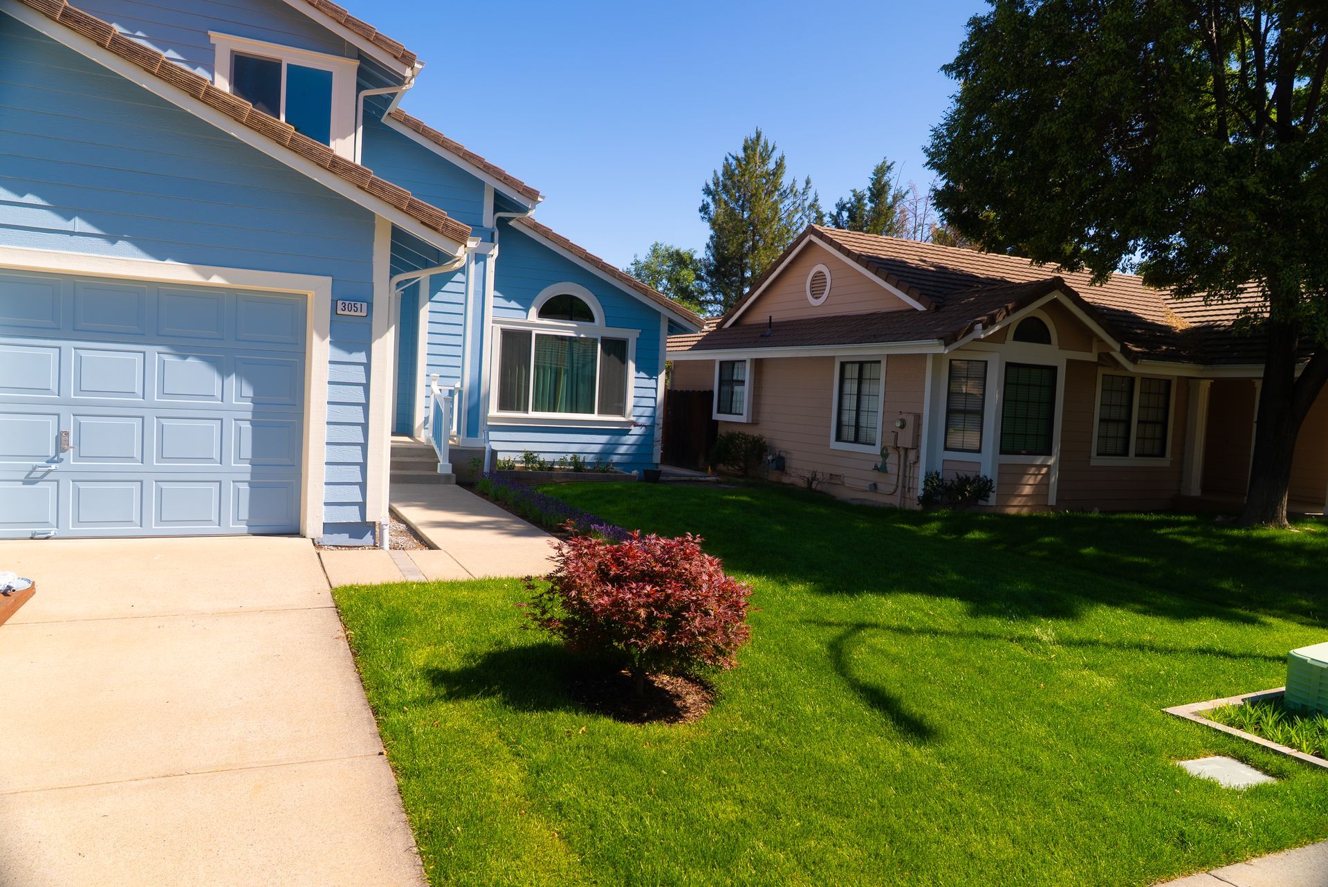 Two-story blue house with a garage next to a smaller, tan house on a sunny day.