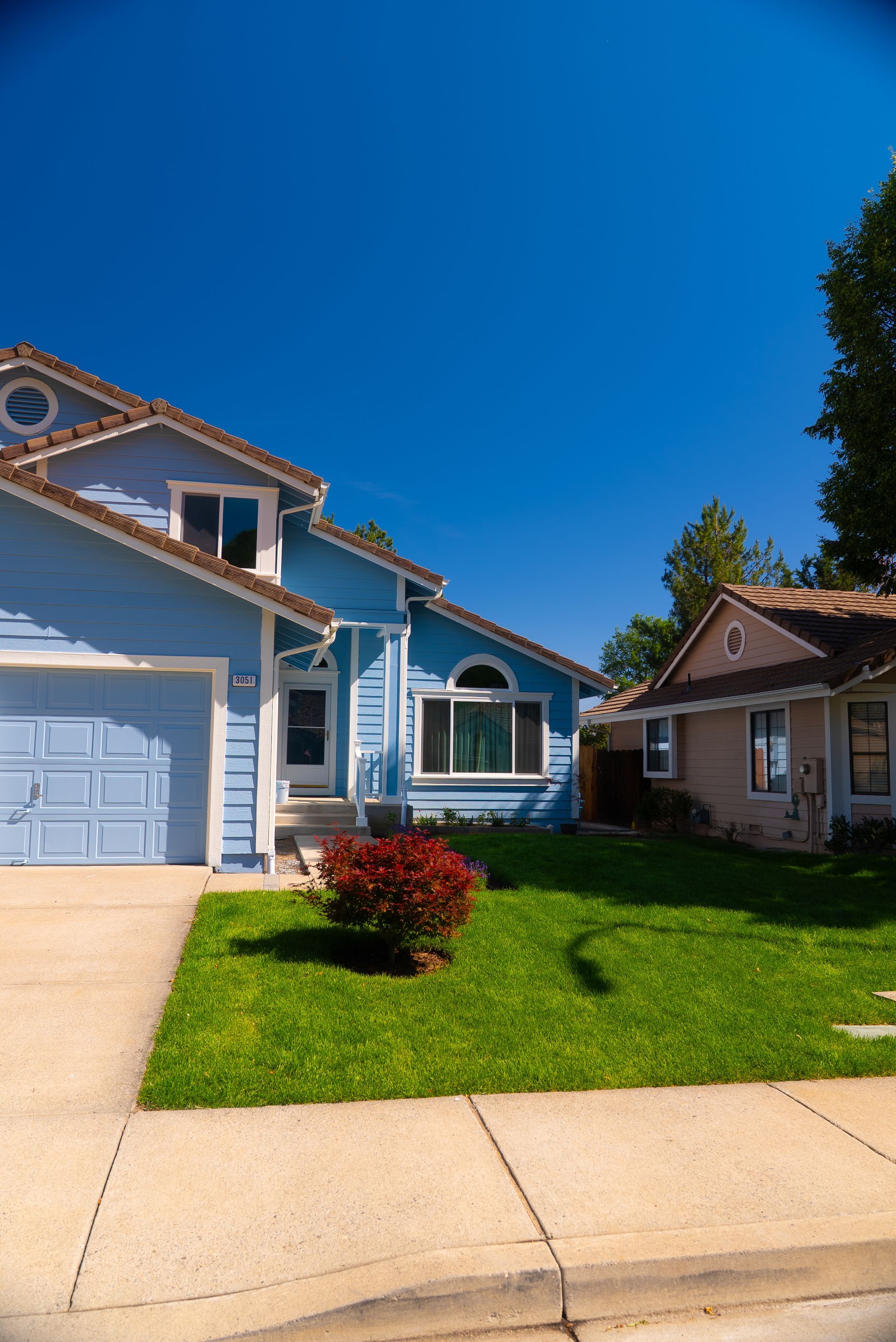 Blue house with garage, green lawn, red bush, bright blue sky, and a smaller house next door.