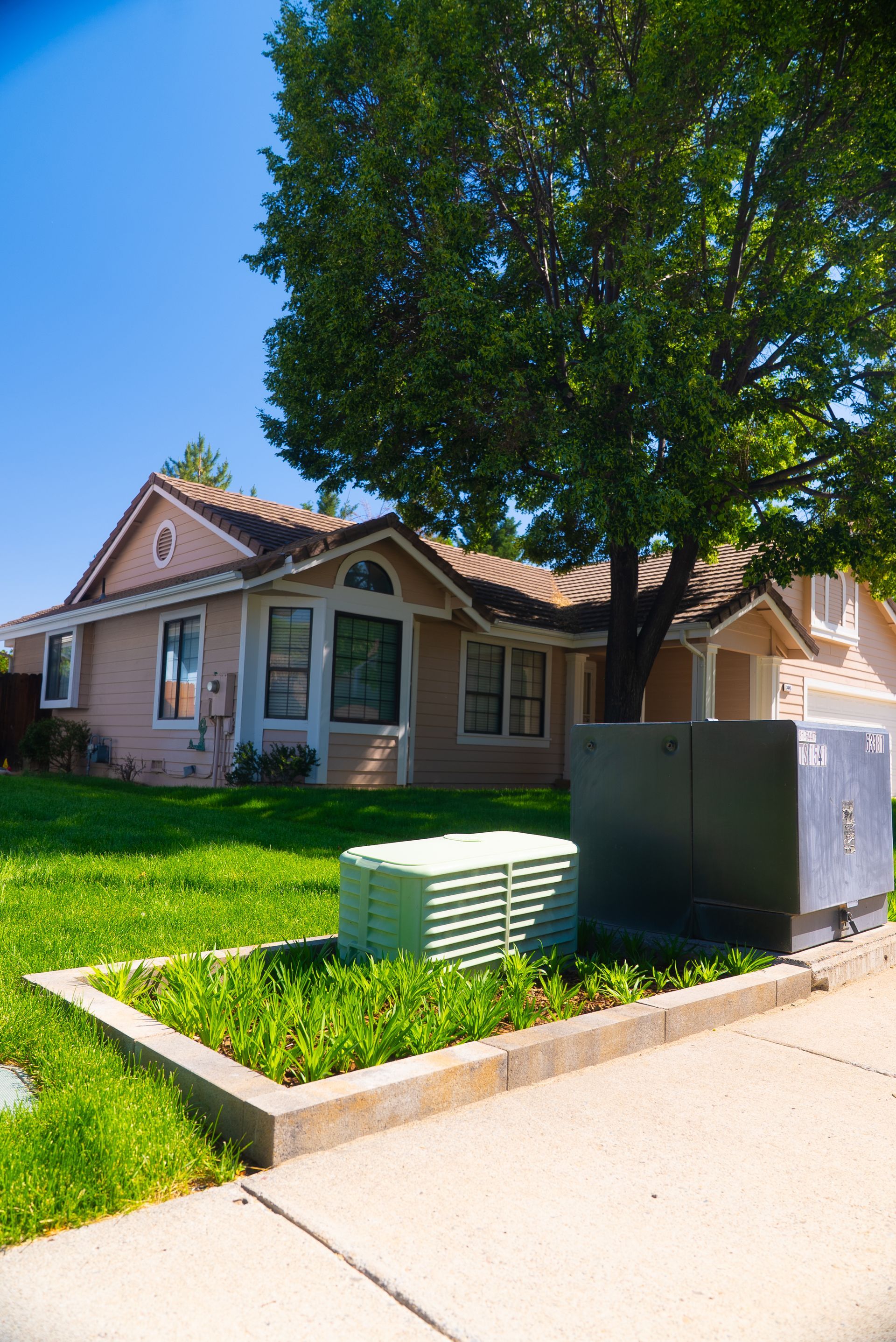 House with tan siding, large tree, and green lawn. Utilities in the foreground, bright blue sky.