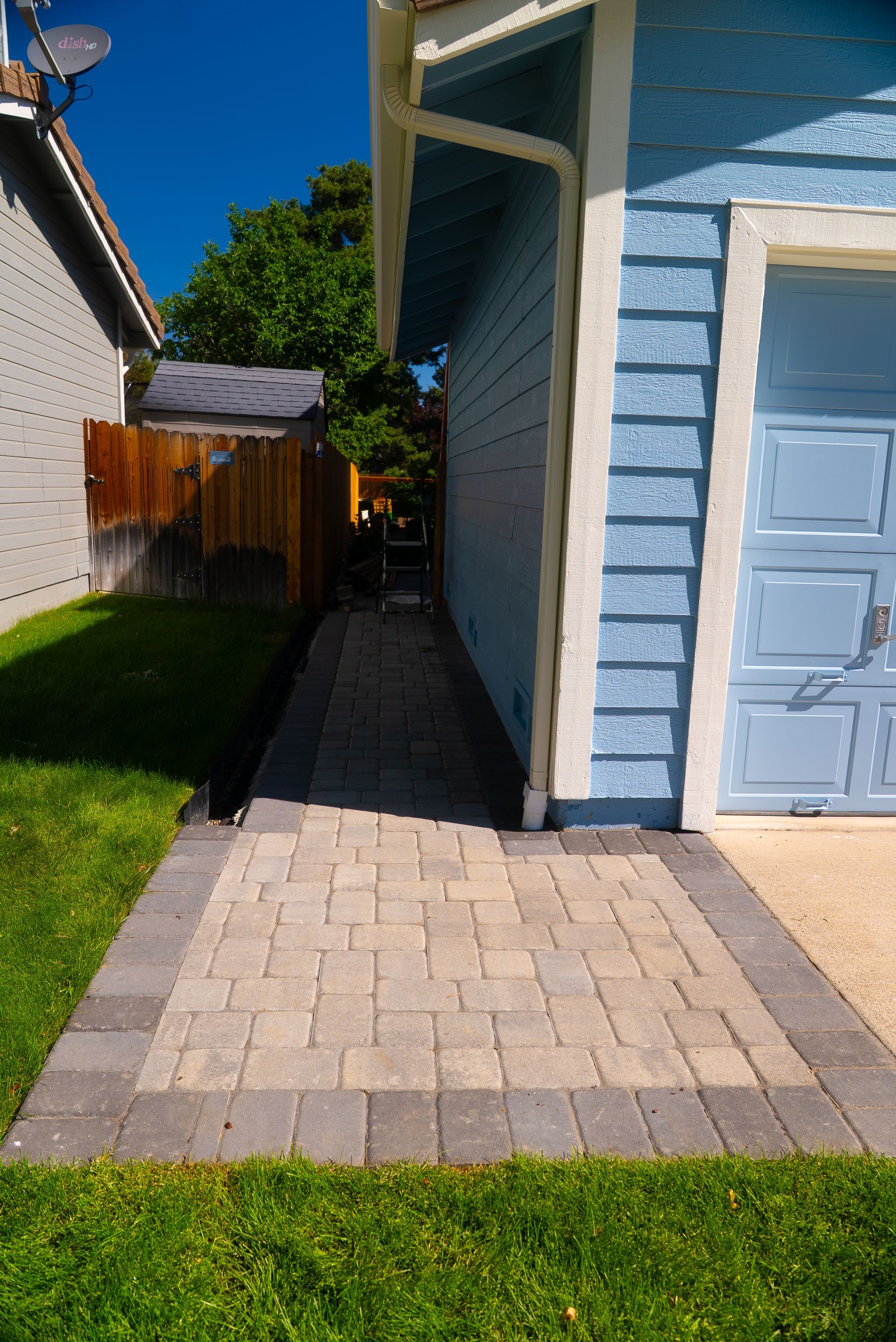 Brick walkway leading alongside a blue house with a garage door. Green lawn on either side.