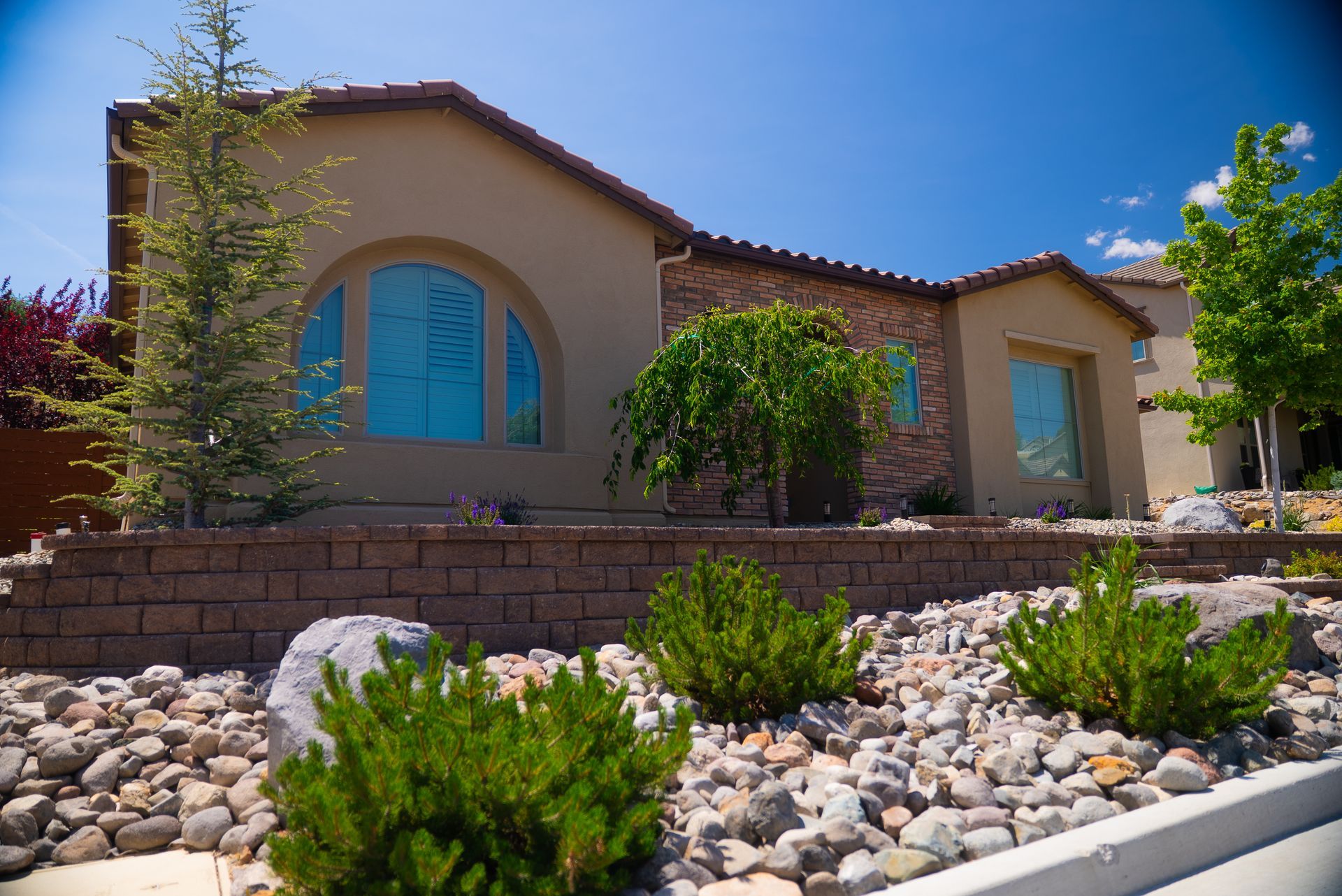 Beige stucco house with stone accent, arched window, and landscaped yard with rock and shrubbery.