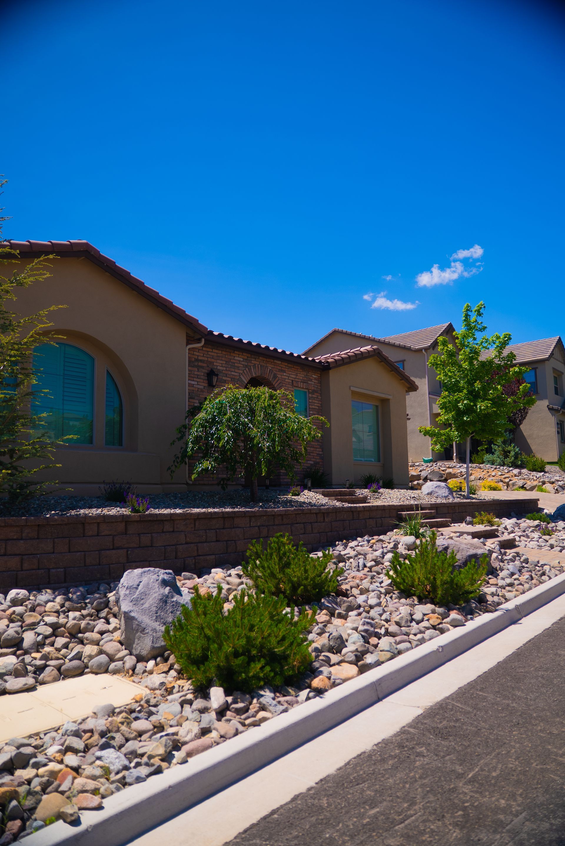 Modern house with beige stucco and stone facade, rock landscaping under bright blue sky.