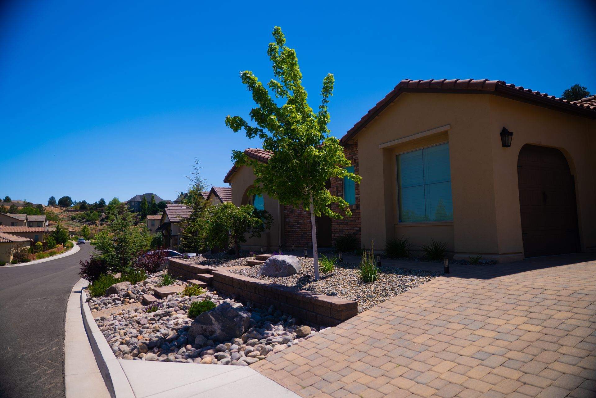 Tan house with a cobblestone driveway, a young tree, and landscaped front yard under a clear blue sky - service by Activescapes.