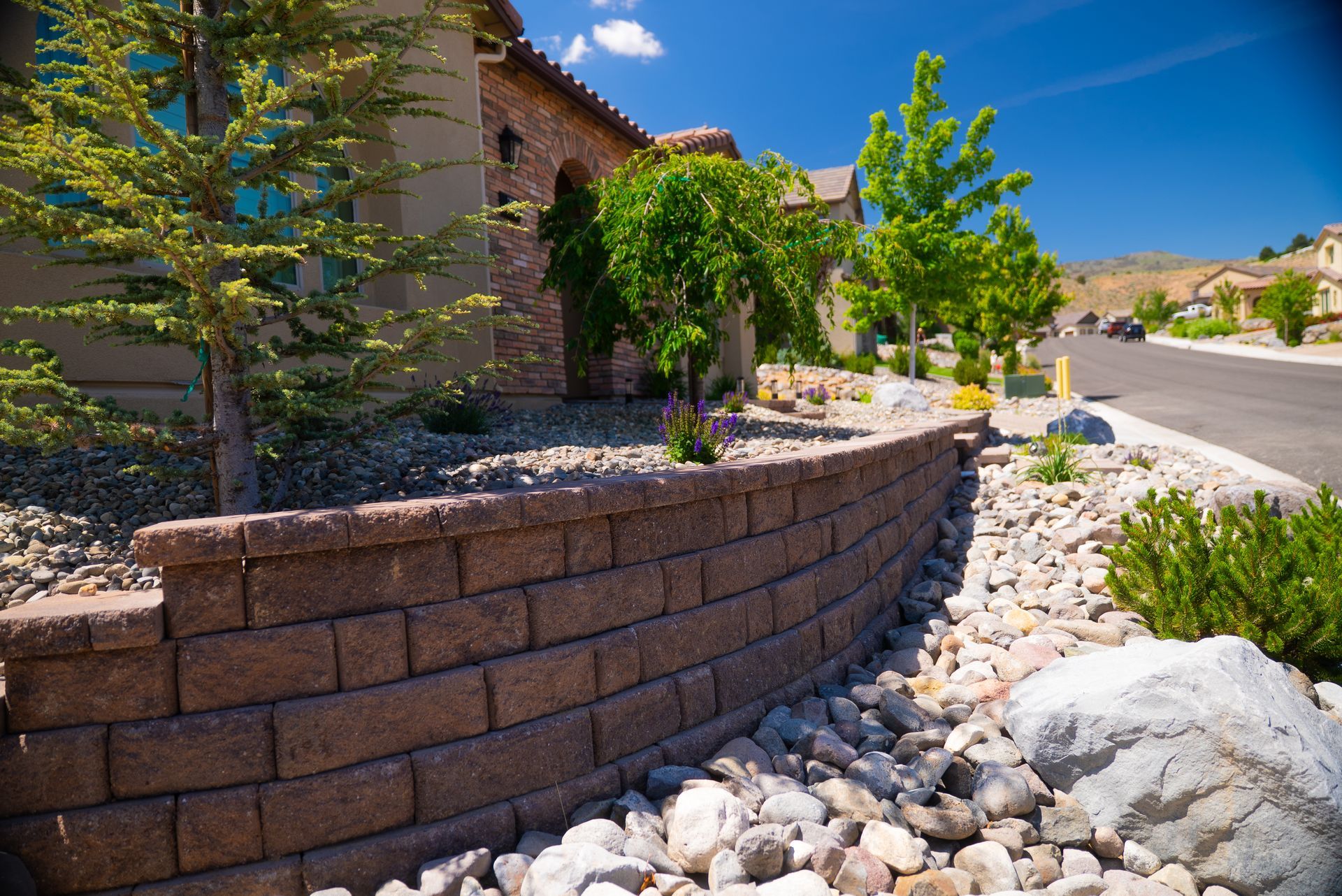 Brick retaining wall in front of a house, landscaping with rocks and small trees under a blue sky.