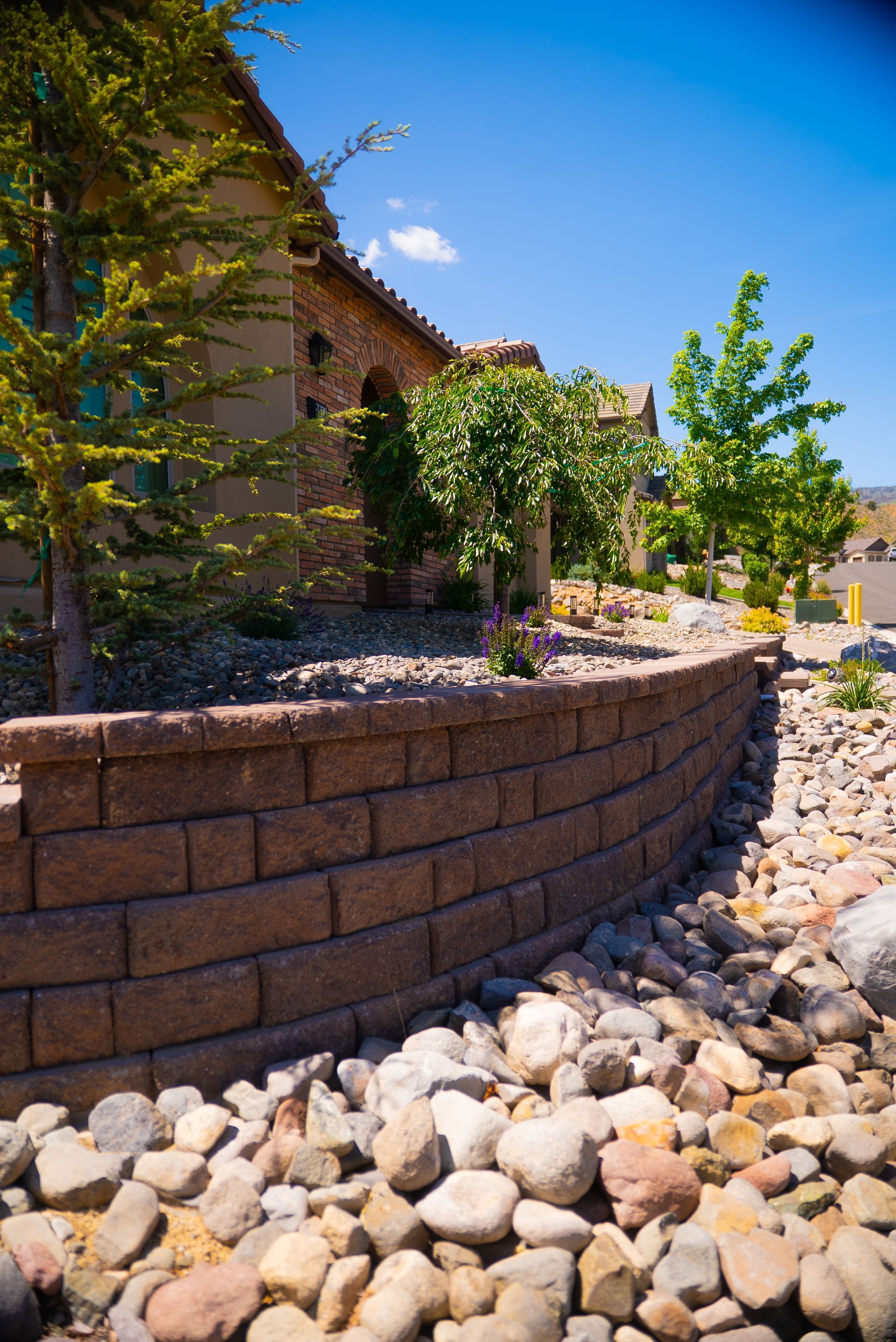 Stone retaining wall with rocks and landscaping in front of a house on a sunny day.