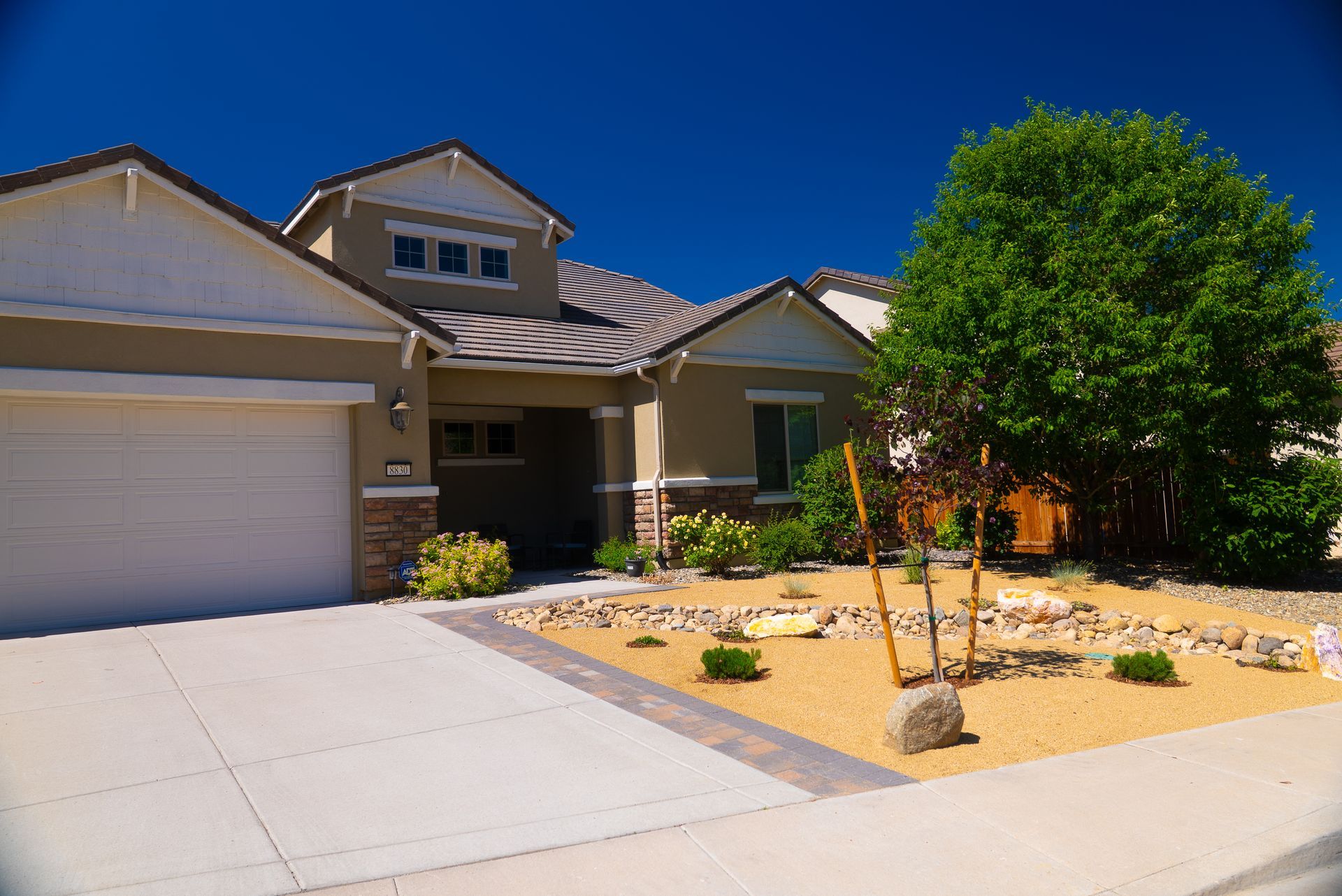 Tan house with white trim, tan and brown landscaping, blue sky.