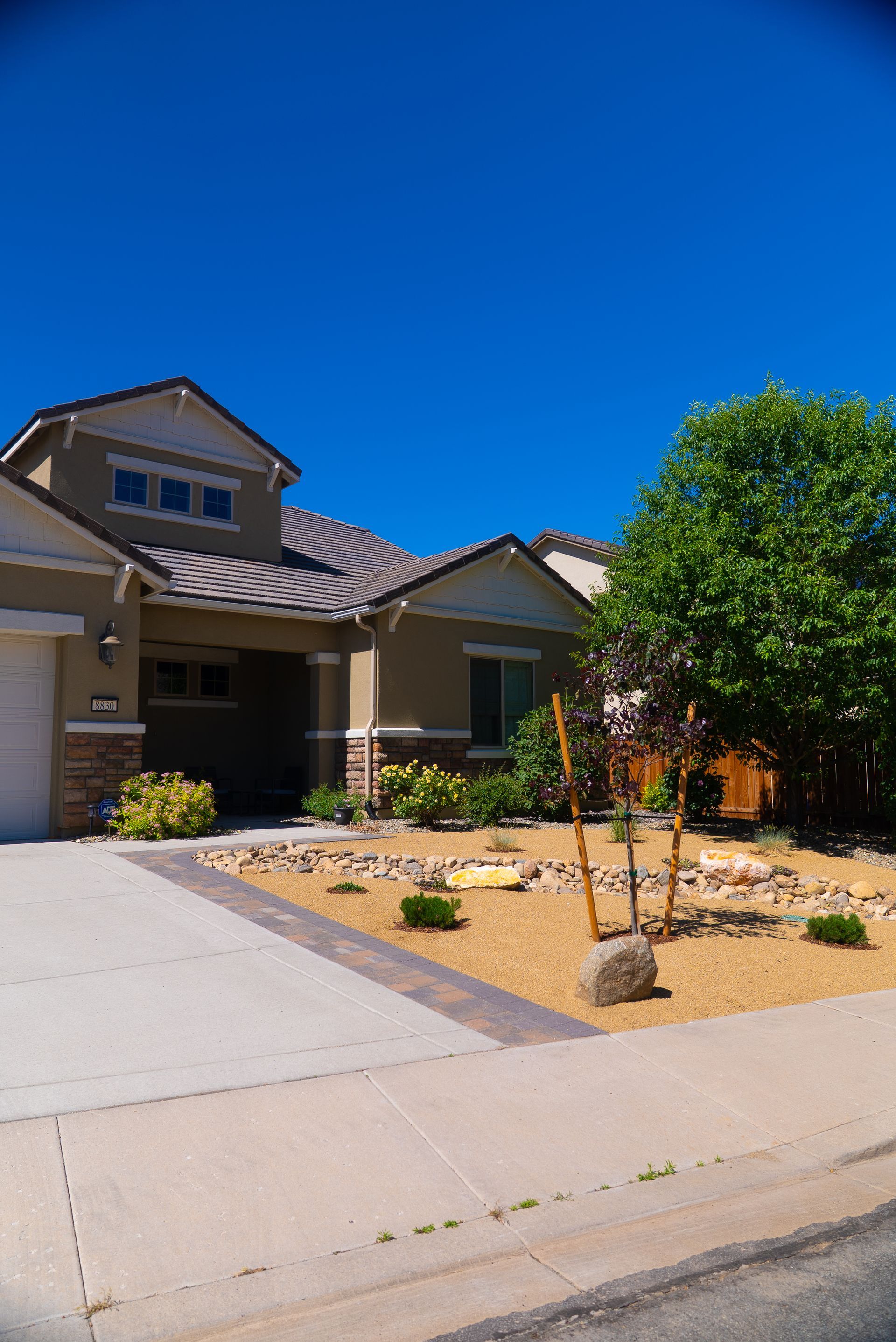 Tan house with a landscaped yard; driveway in the foreground. Blue sky overhead.