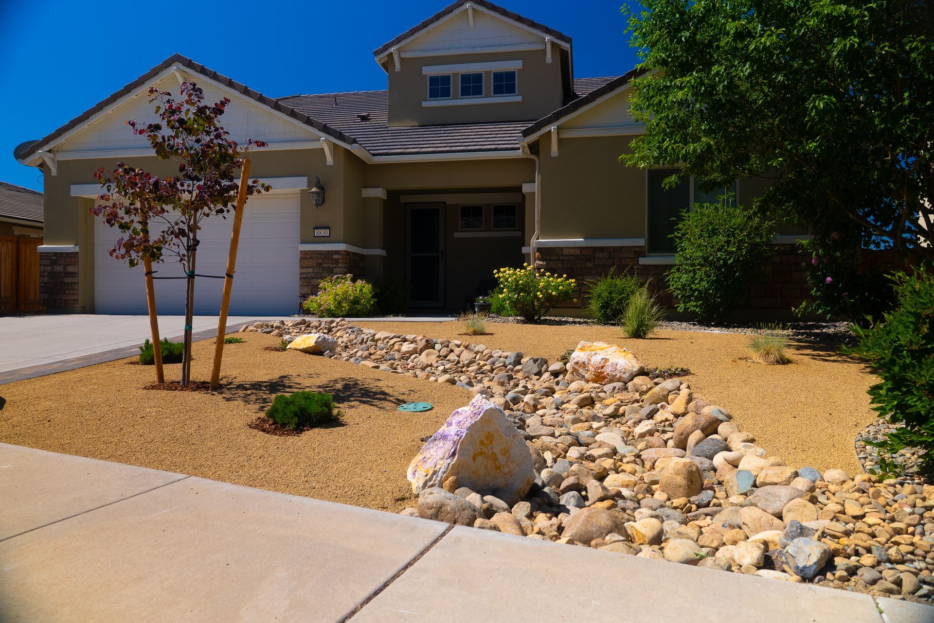 Tan house with rock-lined landscaping, including dry creek bed and large boulder on sunny day.