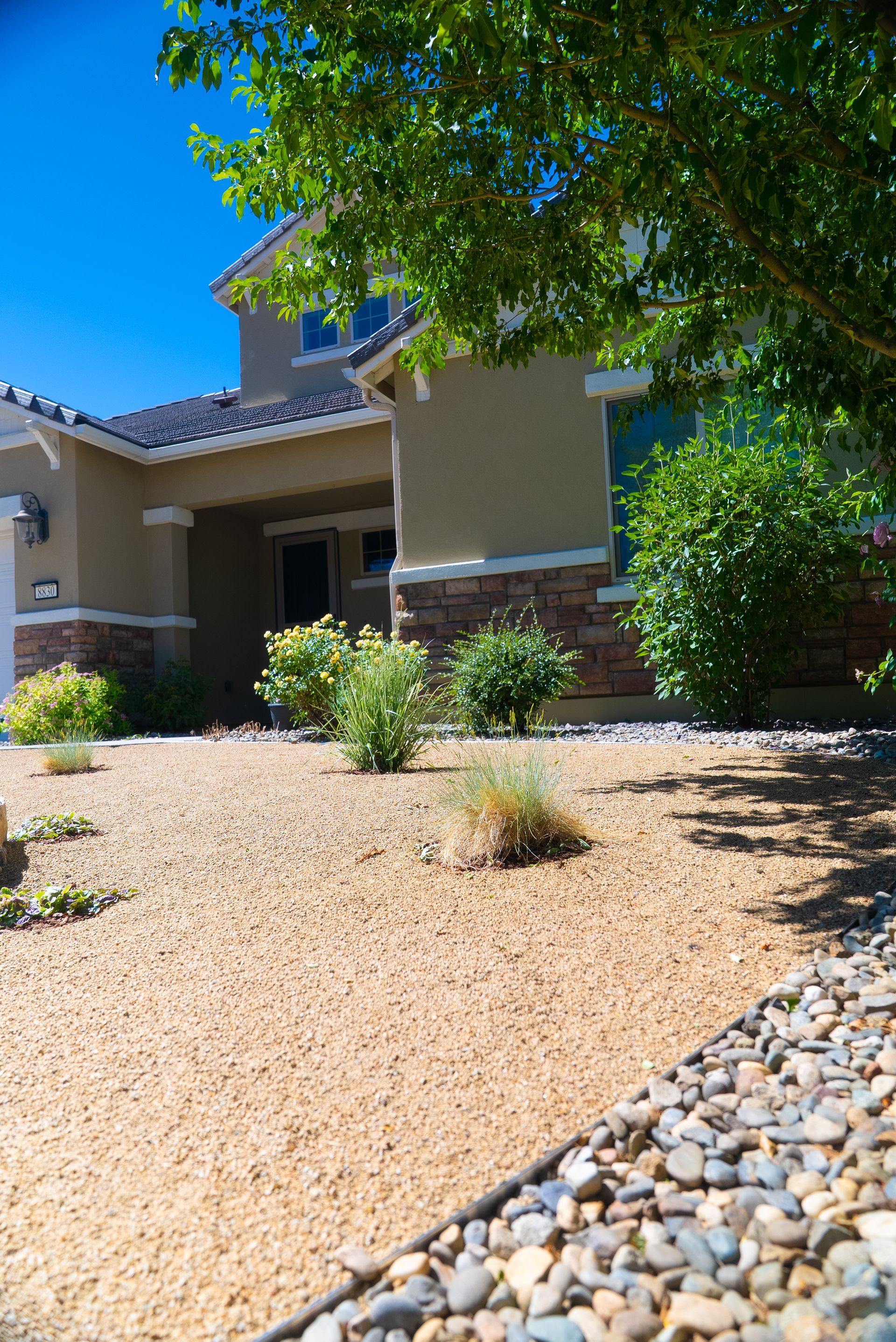 Beige house with a gravel front yard, greenery, and a blue sky.