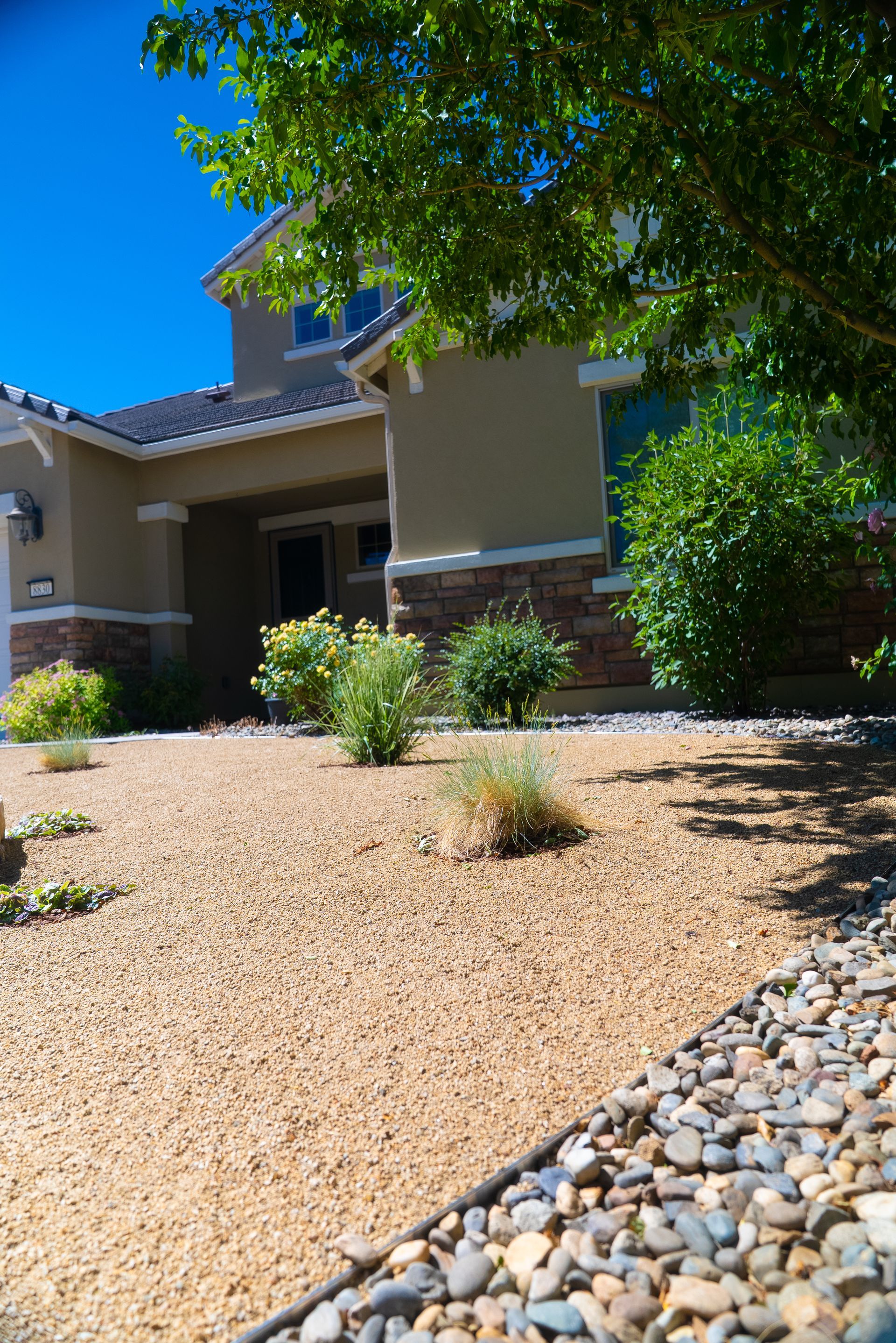 Tan house with gravel yard and sparse greenery under a bright blue sky.