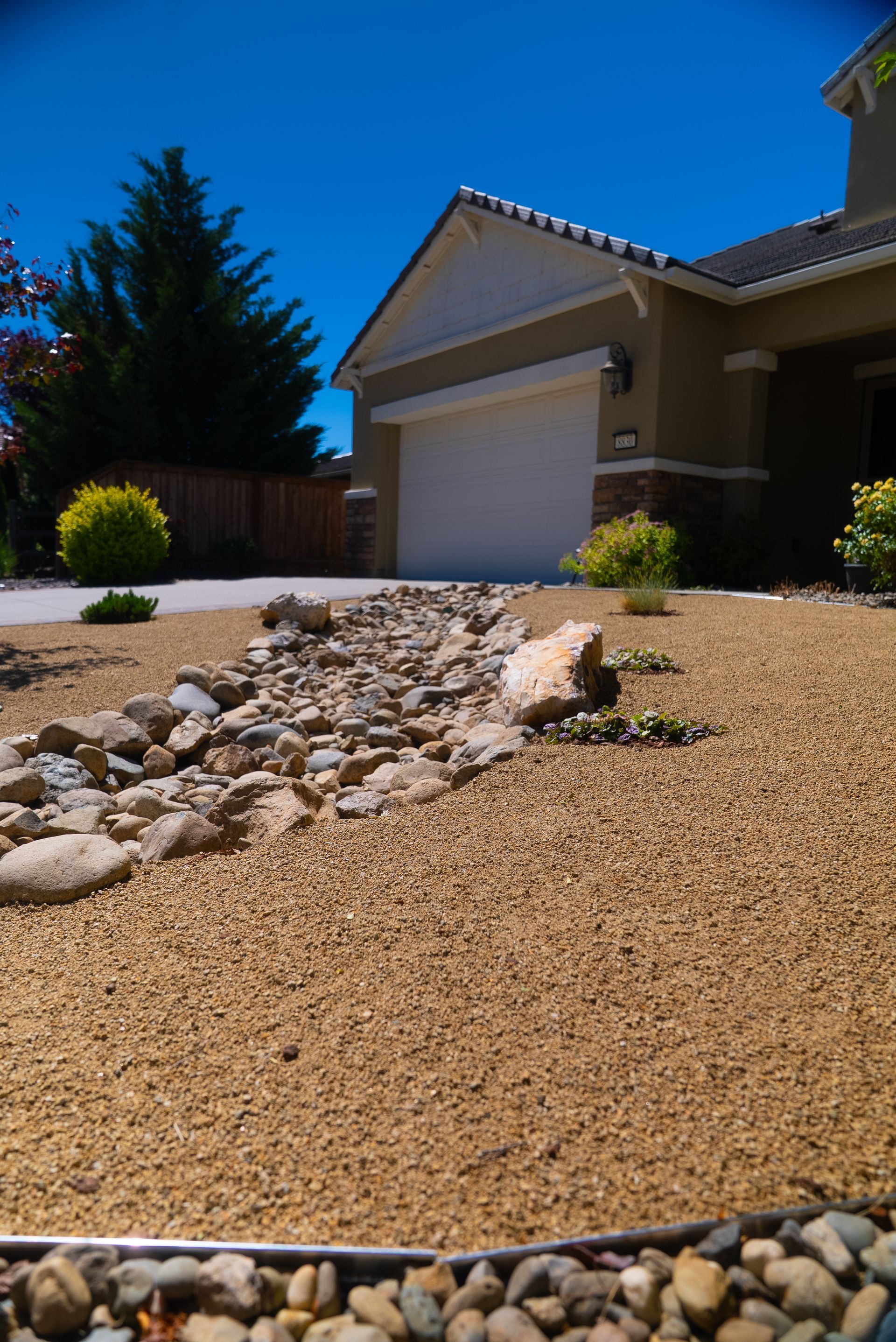 A home's front yard features a dry creek bed with tan gravel and a bright blue sky.