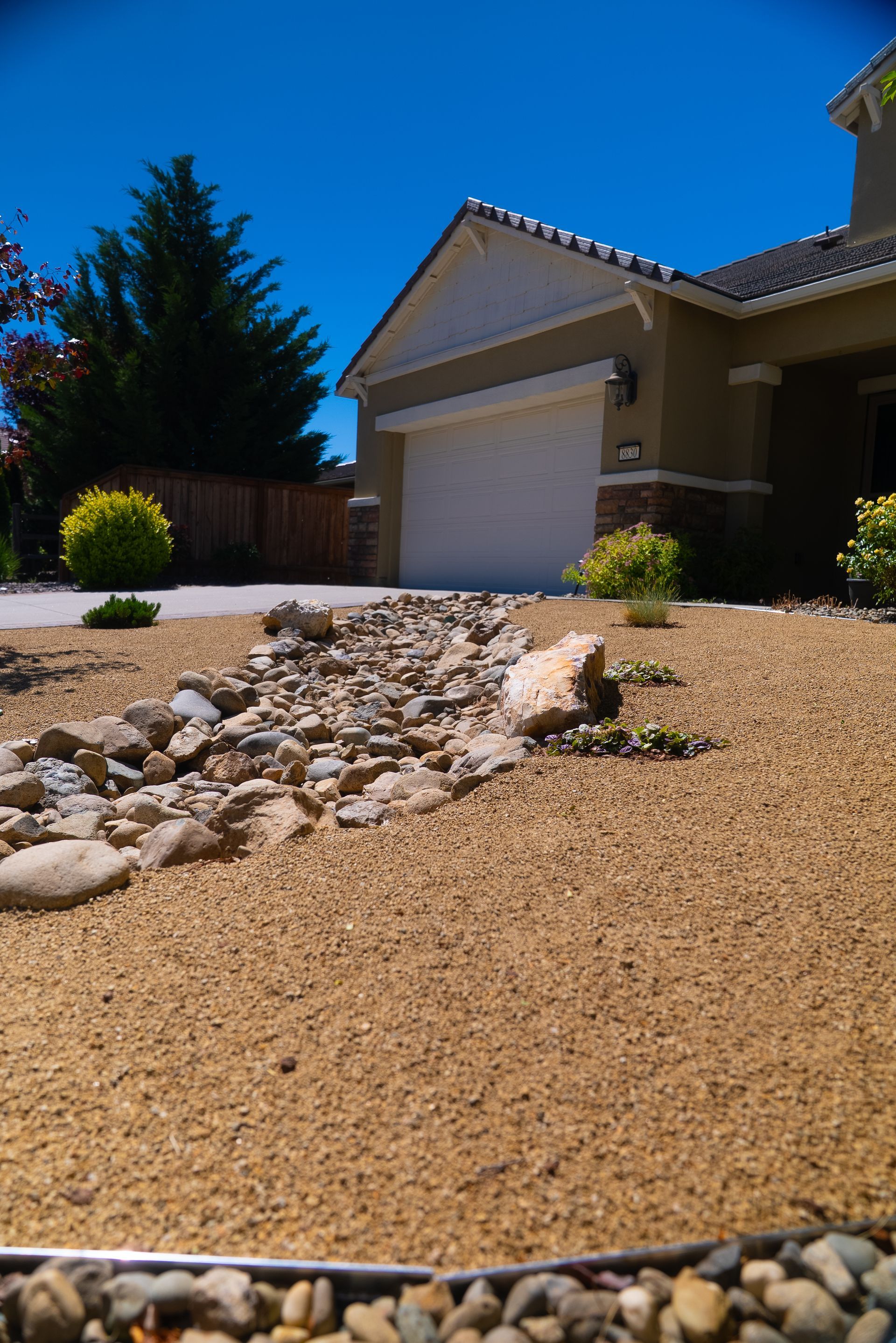 A house with a beige facade, tan gravel yard, and rock-lined dry creek bed under a blue sky.