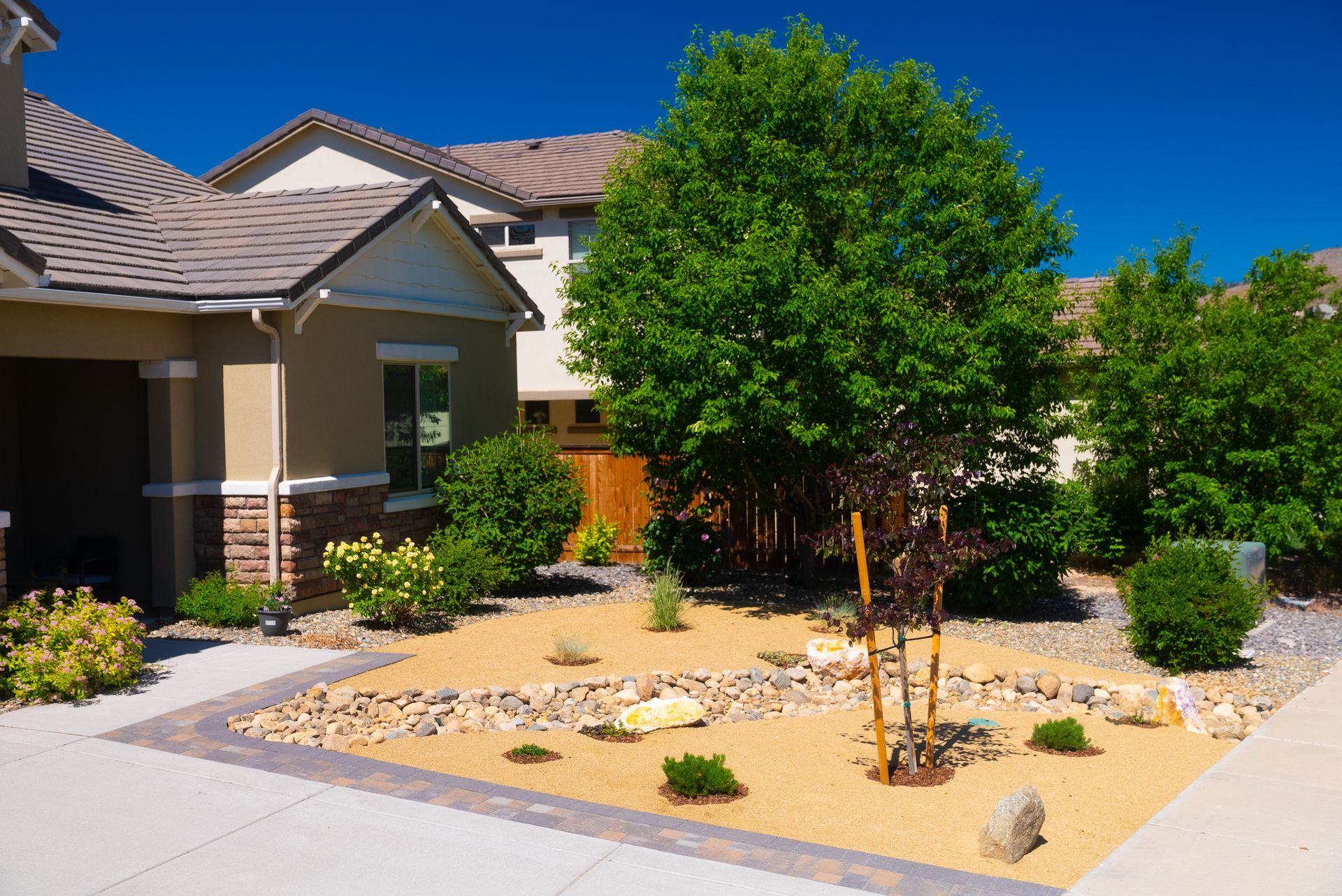 Front yard landscaping with a rock garden, shrubs, and a tree, next to a house with beige siding and a blue sky.