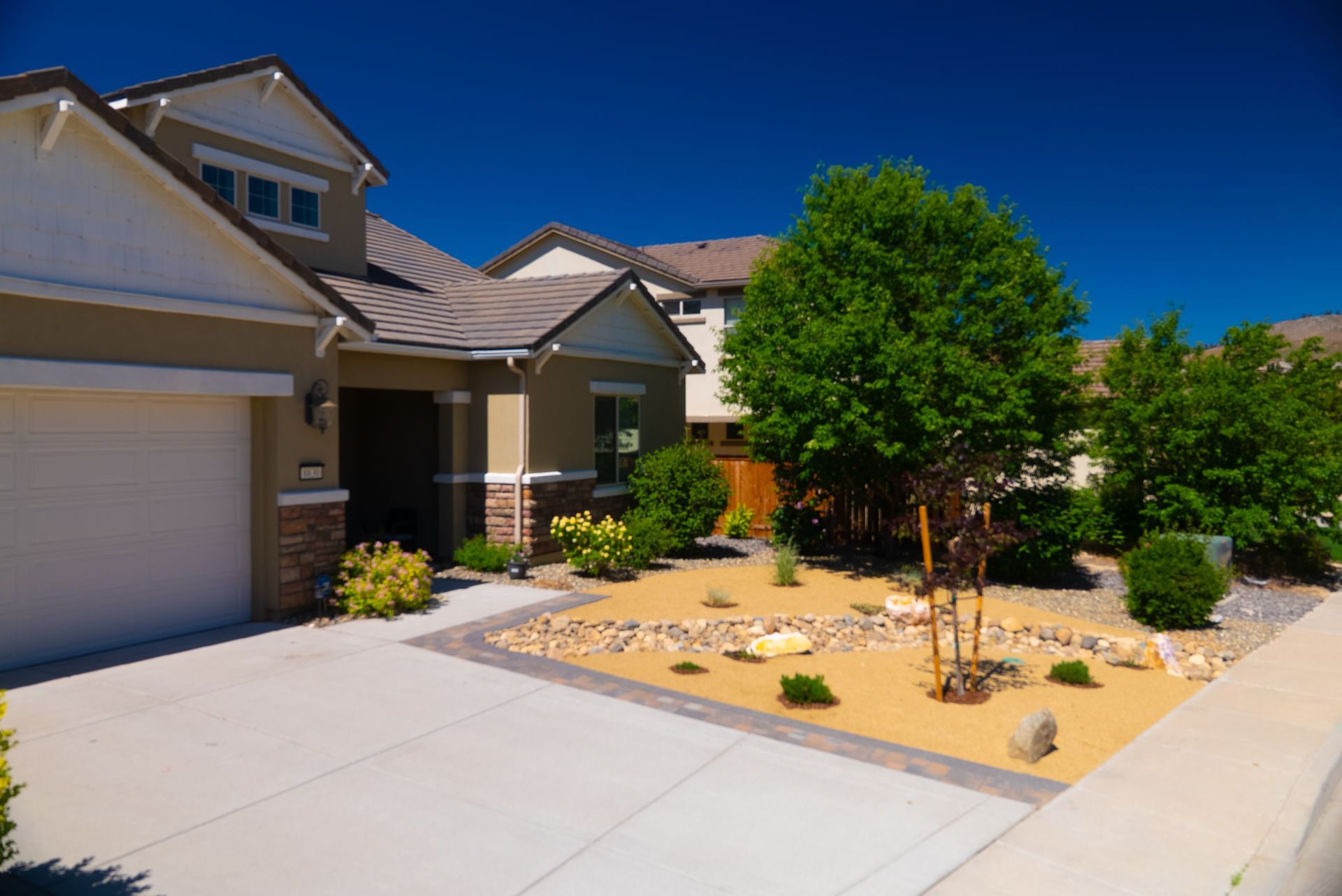 House with tan exterior, driveway, and landscaped front yard with tan gravel and green plants.