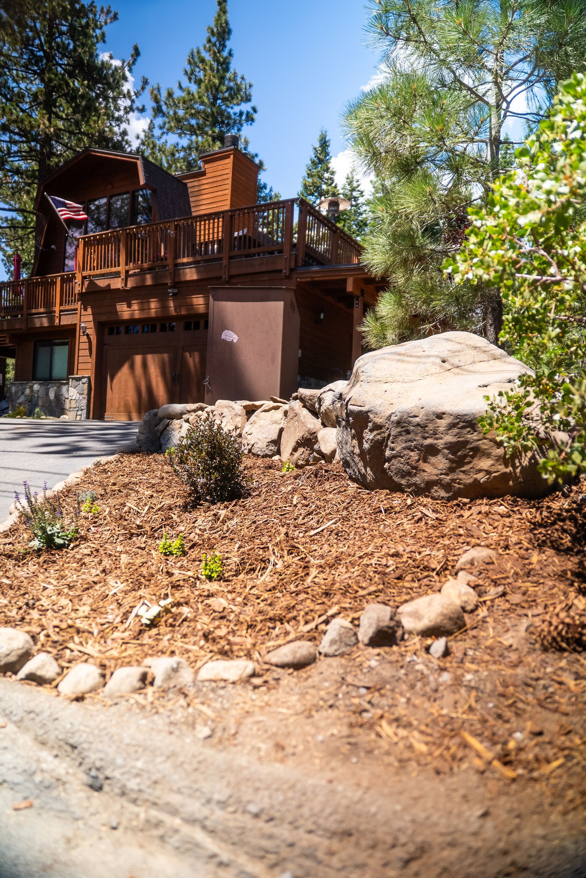 Brown wooden cabin with a deck, driveway, and landscaping on a sunny day.