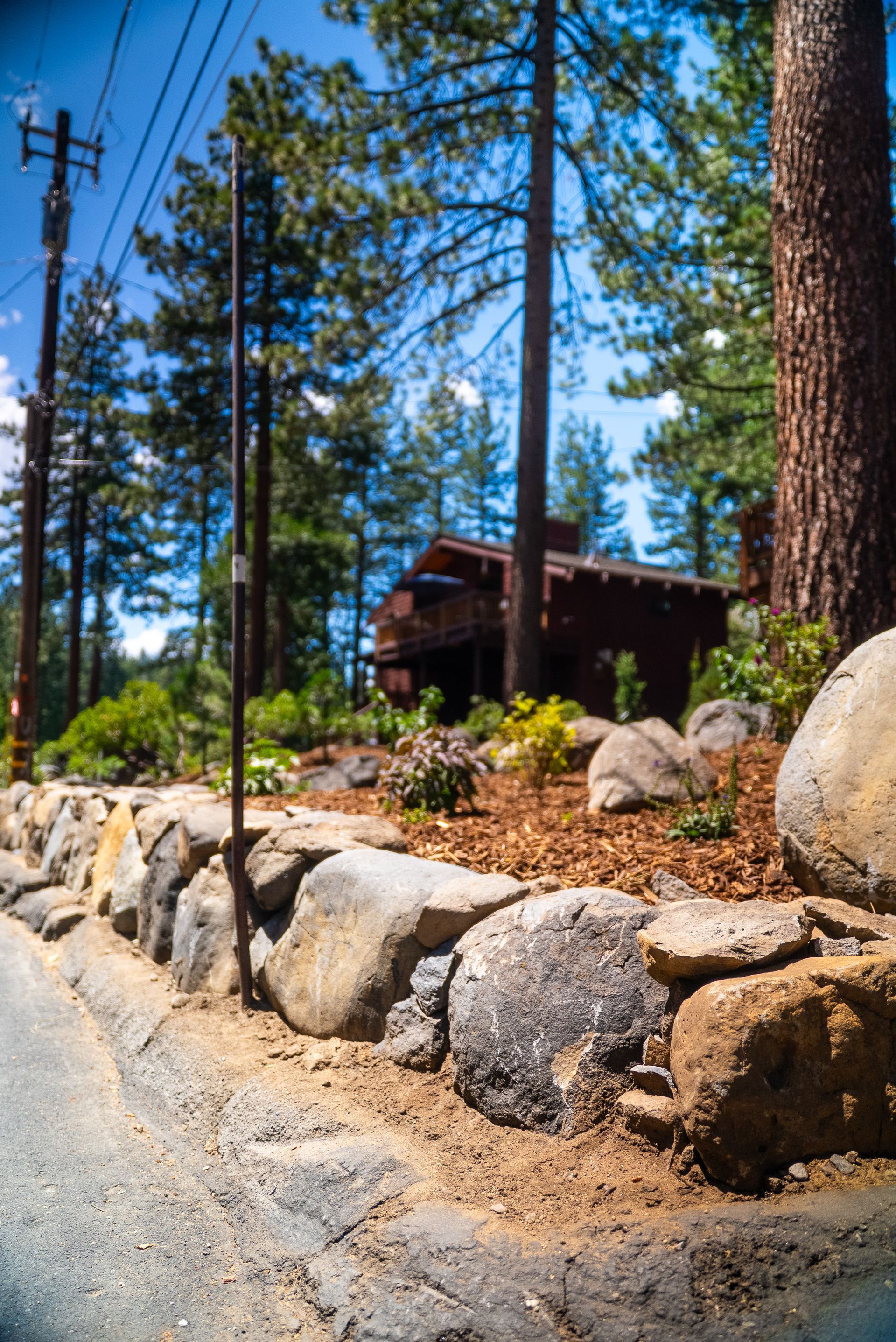 Stone wall borders a road. A brown cabin sits behind trees under a blue sky.